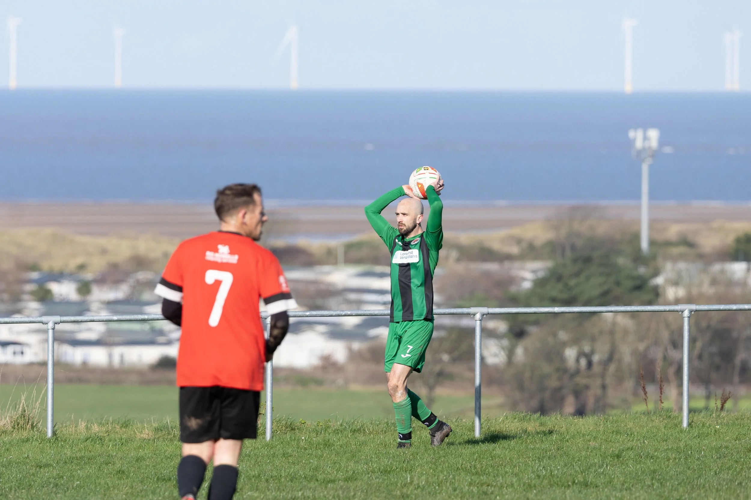 A soccer player in a green uniform holding a ball above his head preparing to throw it in, with another player in a red jersey numbered 7 standing nearby on a grassy field in front of a backdrop of wind turbines and water.