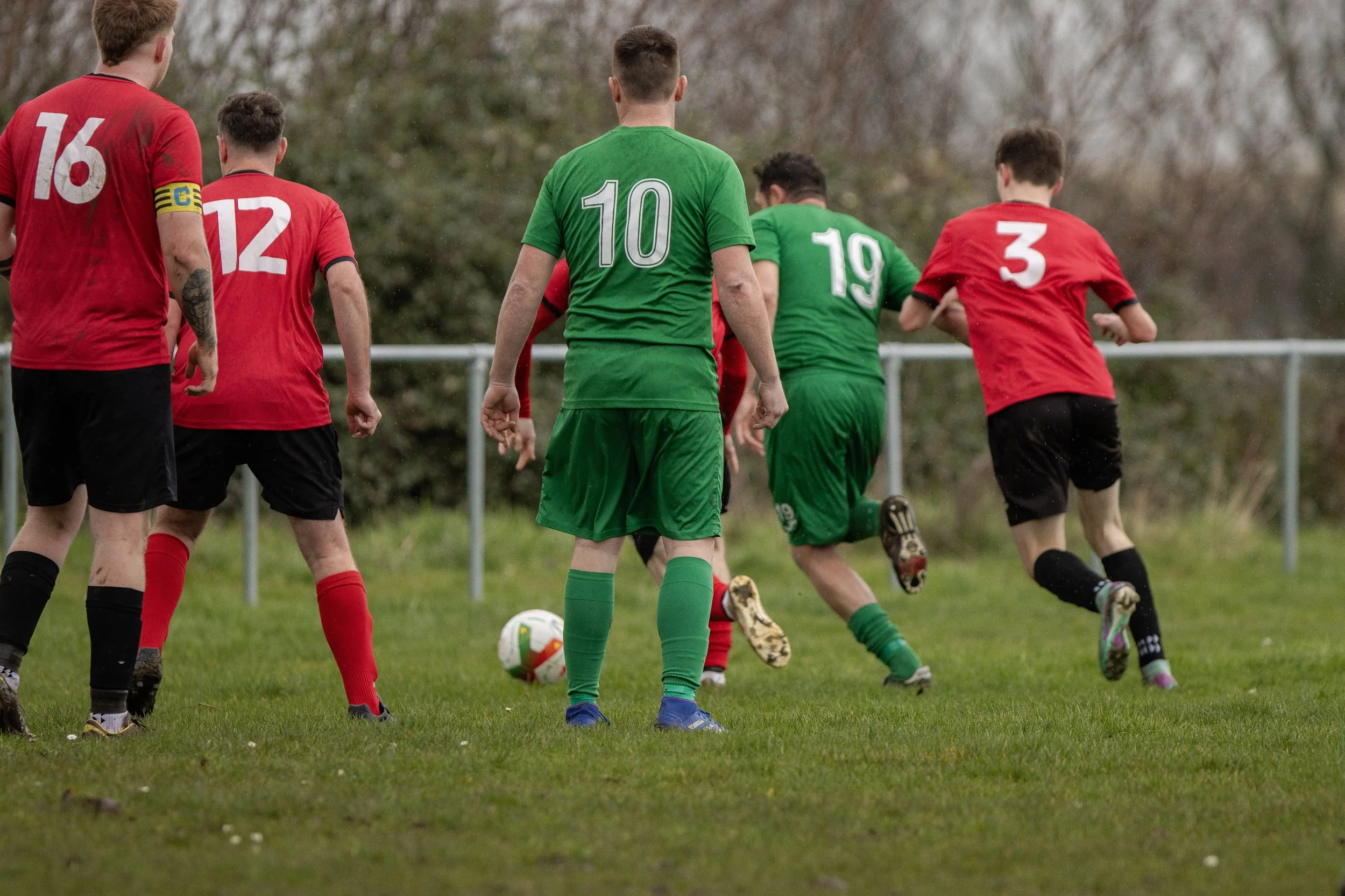 A group of boys playing soccer on a grassy field, wearing red and green jerseys with numbers, with some running towards a soccer ball.
