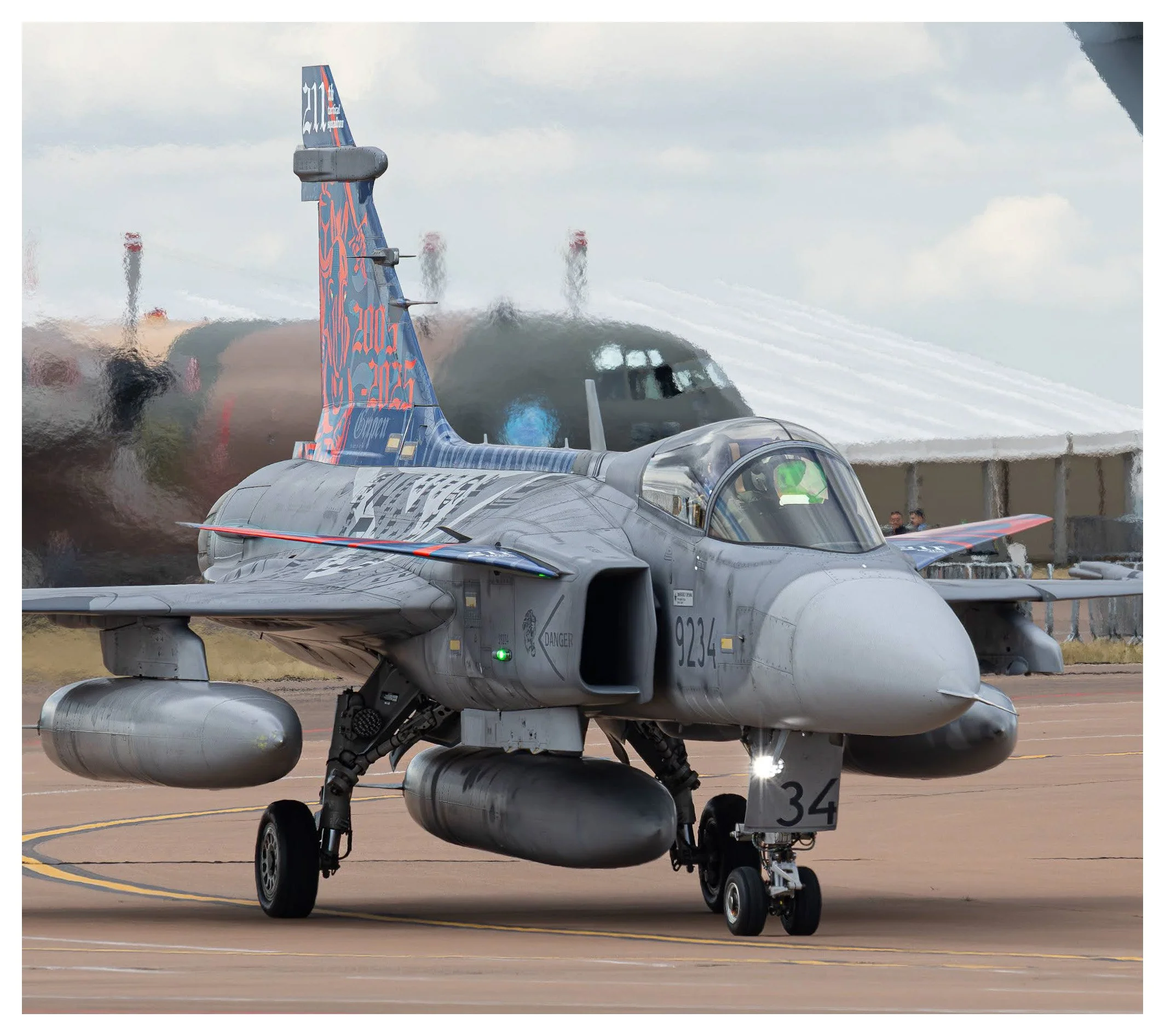 A military fighter jet on the tarmac with a tent structure and another aircraft in the background at an airshow.
