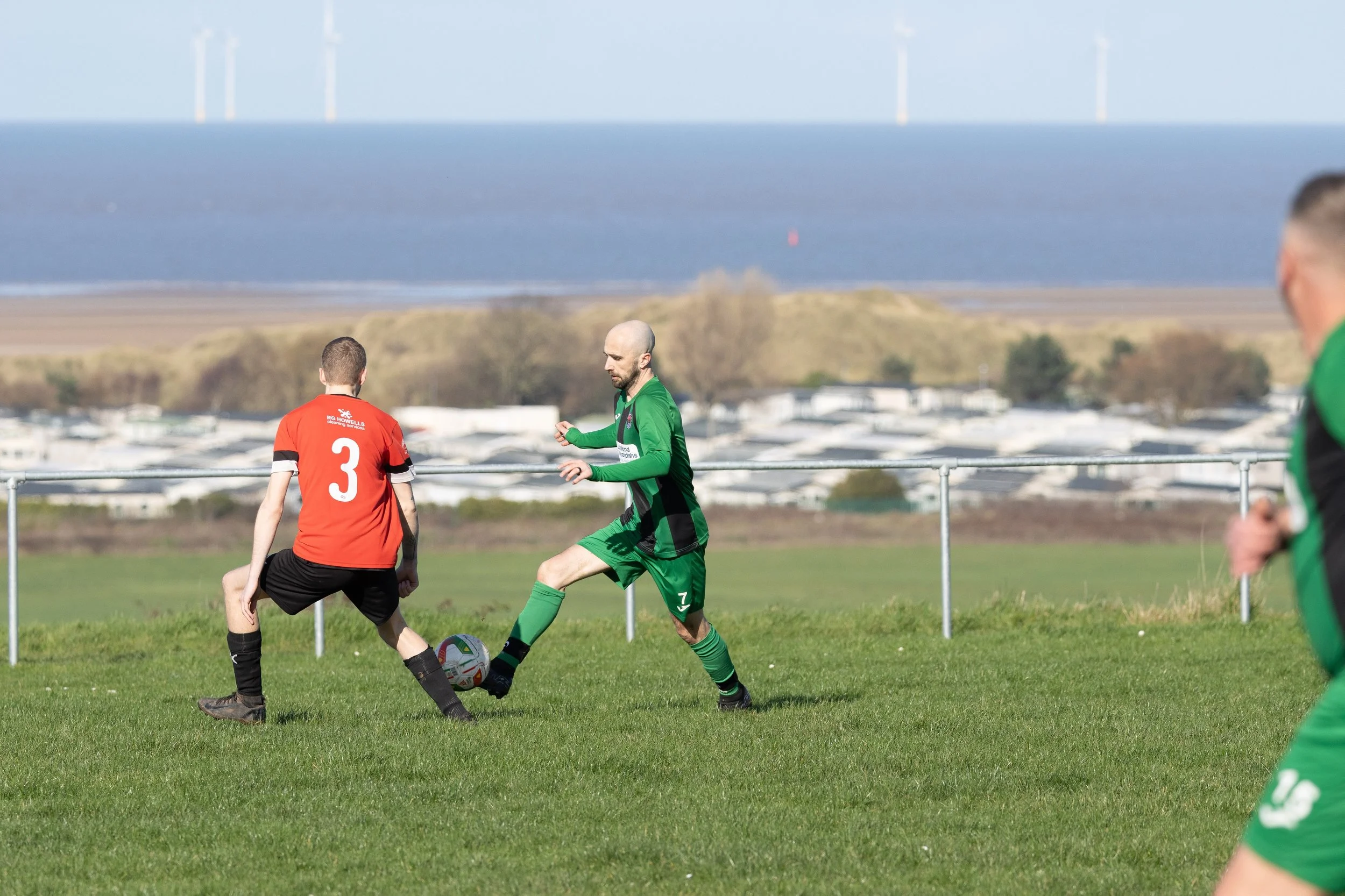 A soccer match with players in red and green jerseys on a grass field near the coast, with wind turbines in the background.