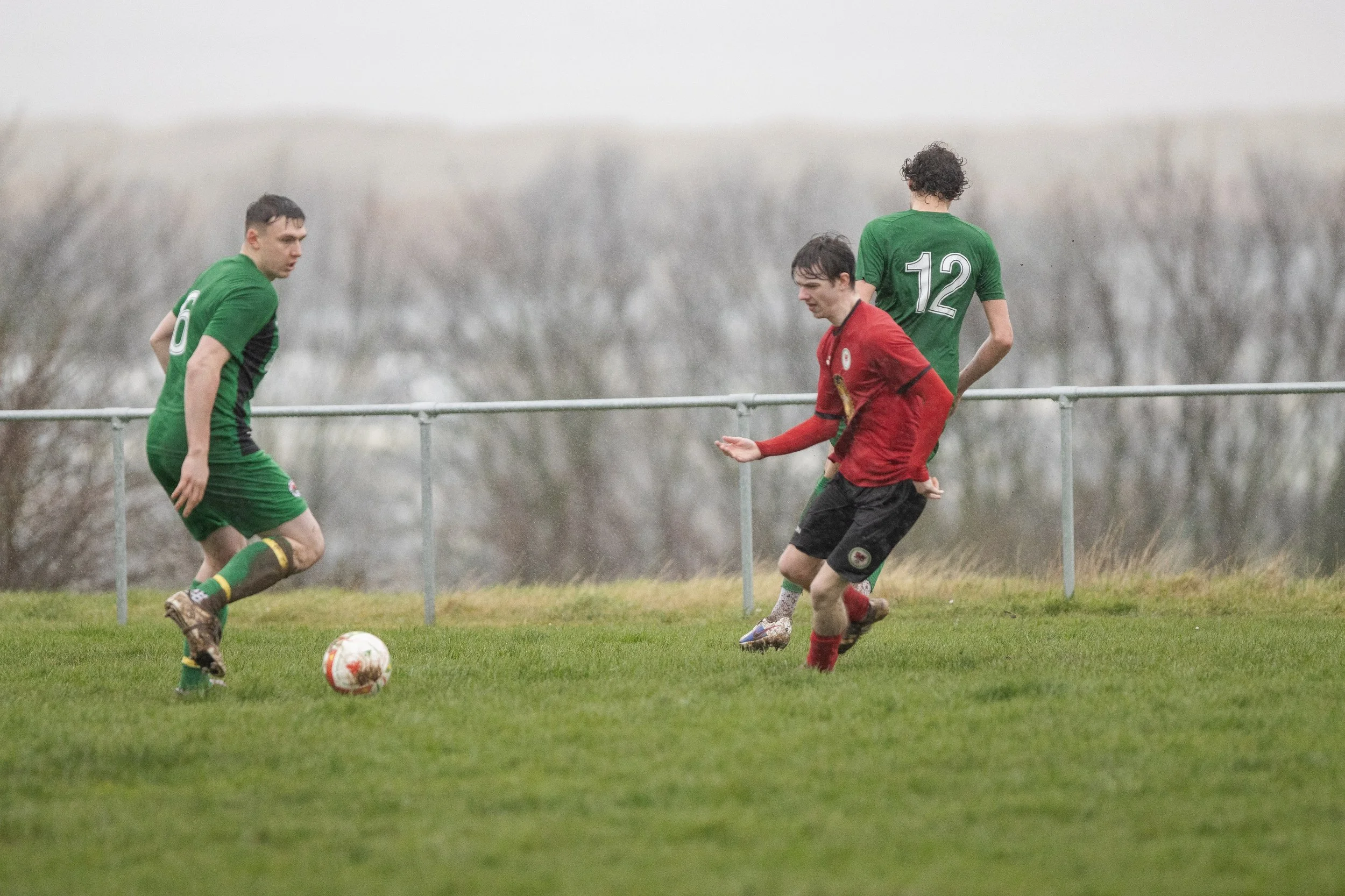 Three soccer players on a grassy field during a match, with two players in green jerseys and one in a red jersey, and a soccer ball in front of them.