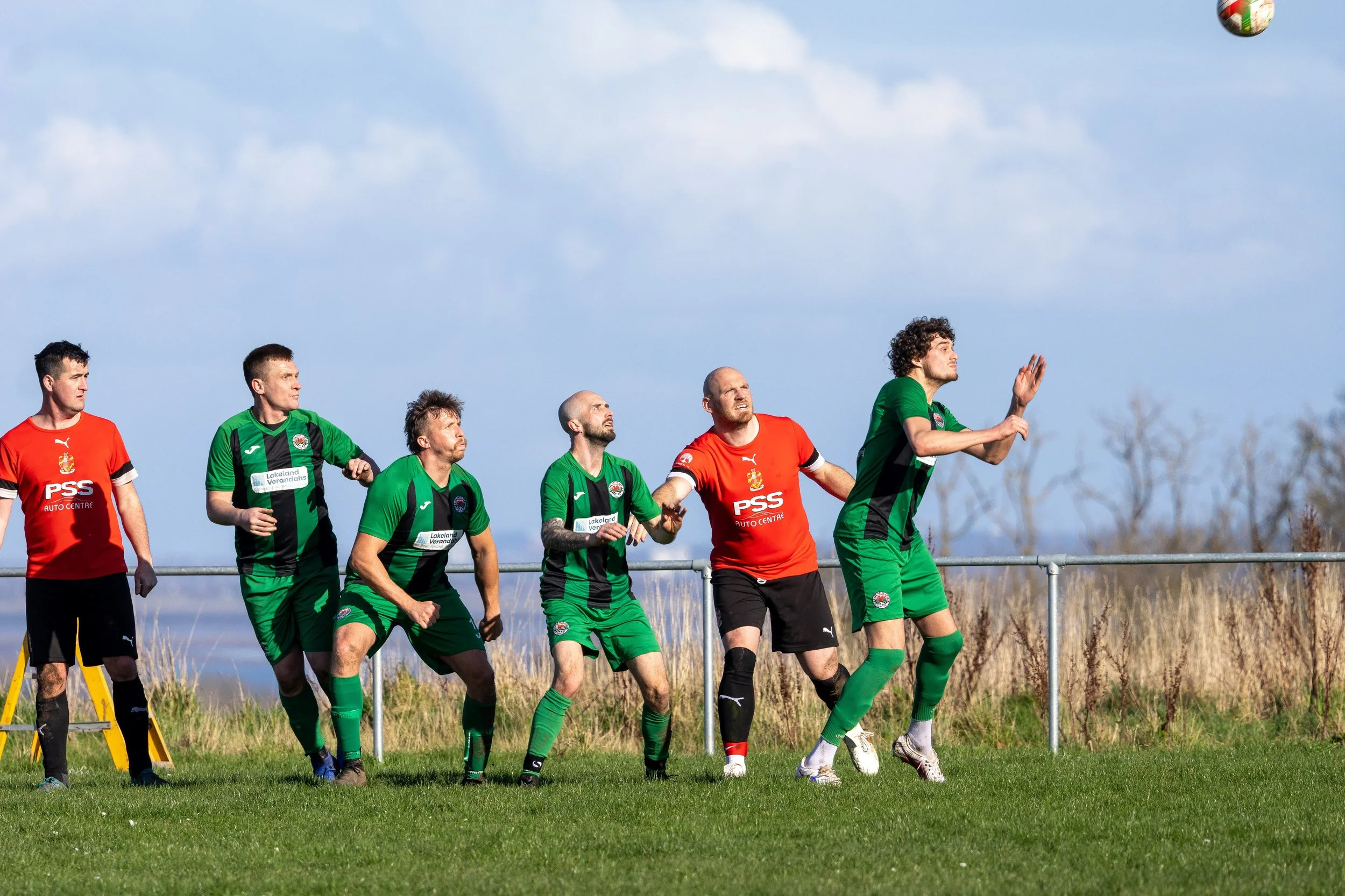 Soccer players in green and red jerseys jumping and preparing to head a ball during a match on a grassy field with blue sky and trees in the background.
