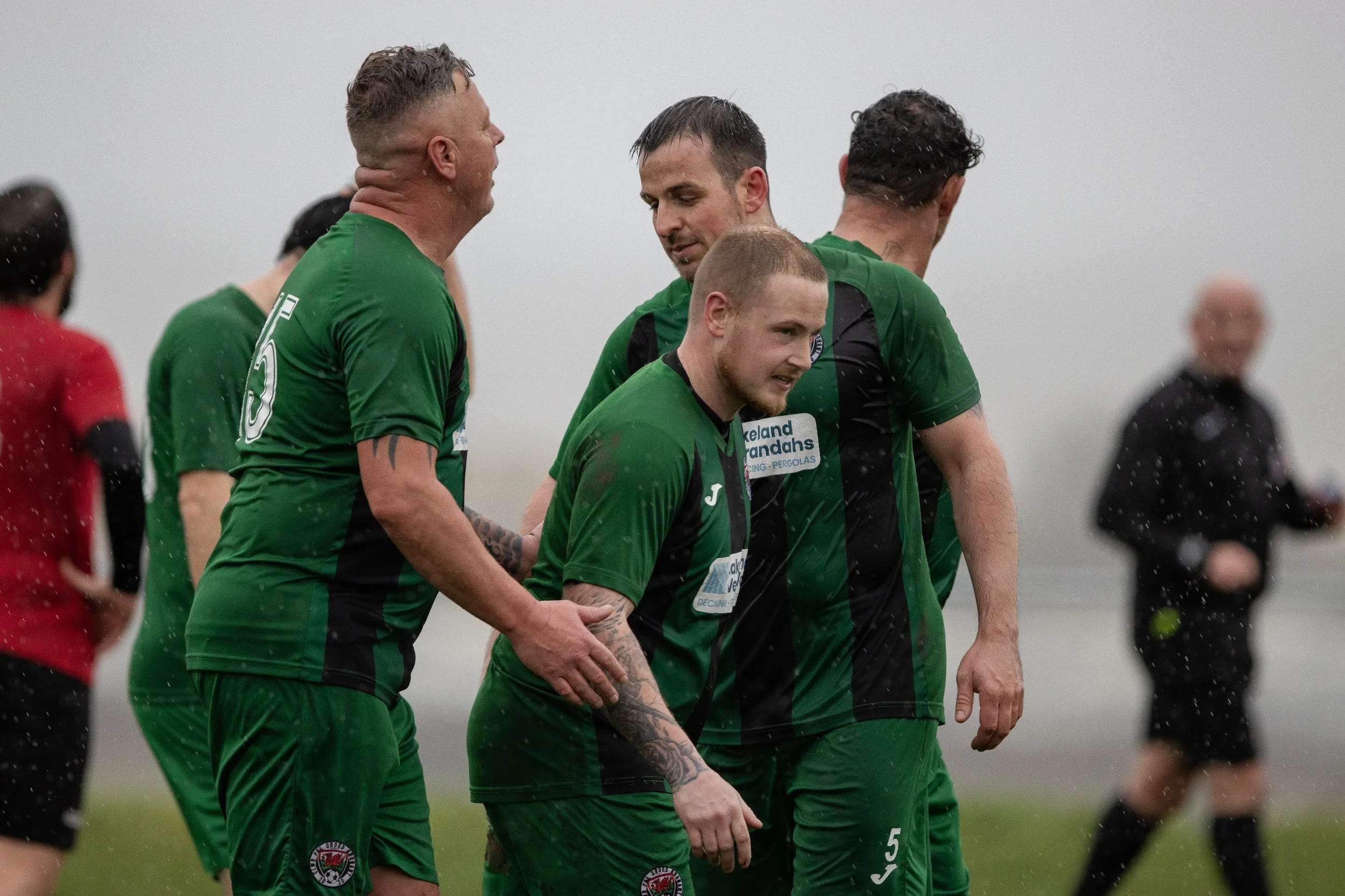 Soccer players in green jerseys celebrating on a field in rainy weather, with a referee in the background.