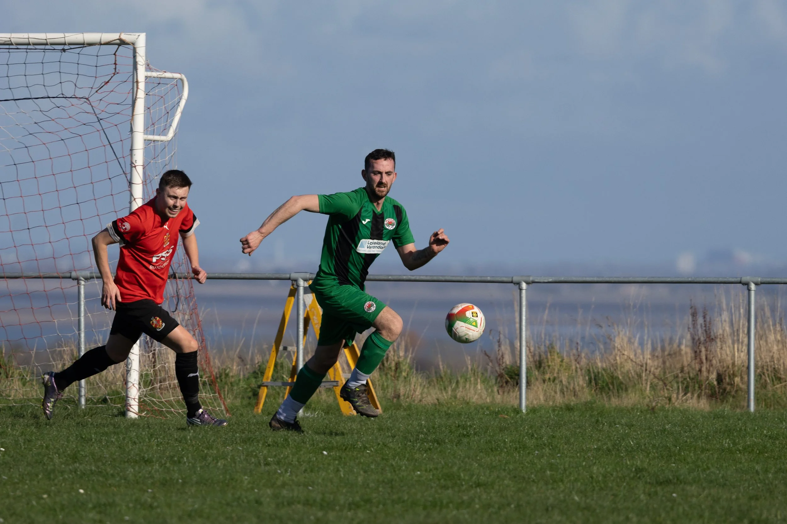 Two soccer players, one in a red jersey and the other in green, are on a field near a goalpost. The player in green is running towards the ball while the player in red follows behind. The background shows a cloudy sky and an open field.