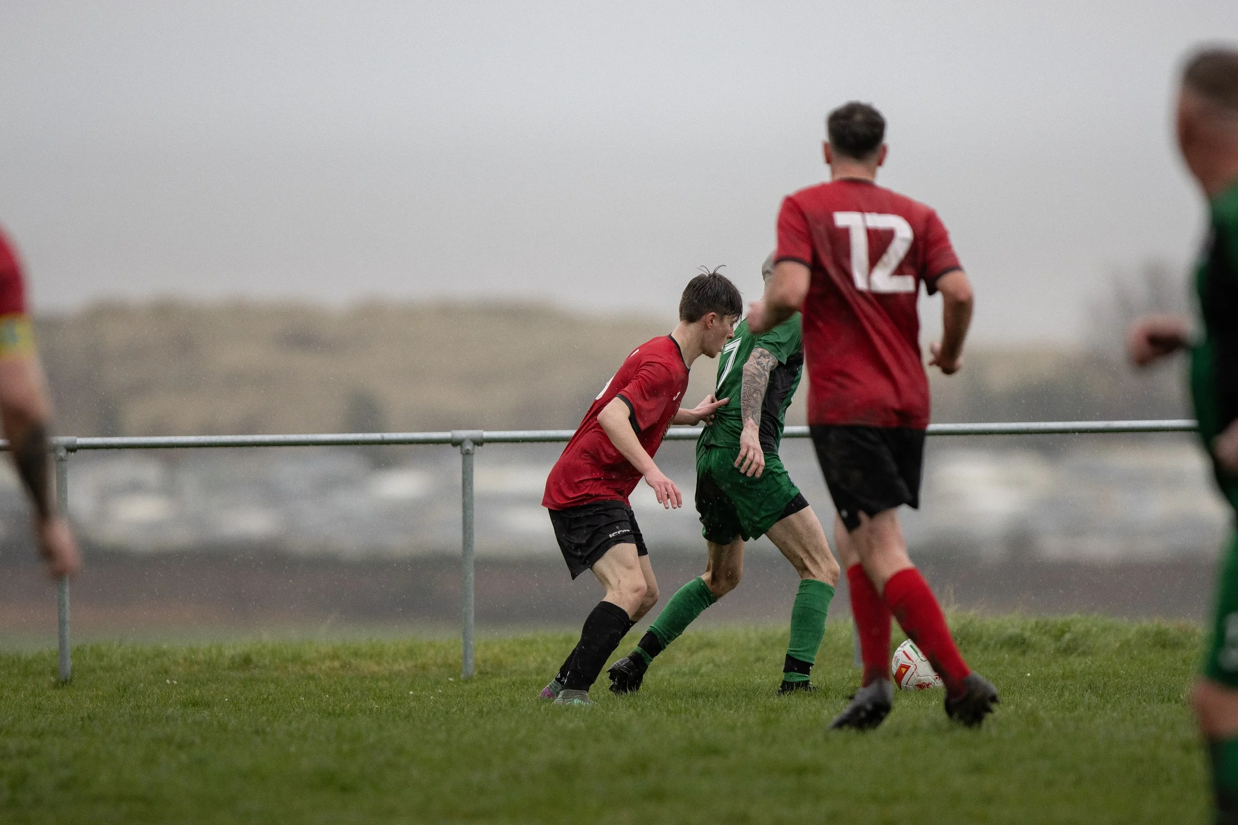 Soccer players on a rainy field, two in red jerseys and two in green, competing for the ball near the fence.