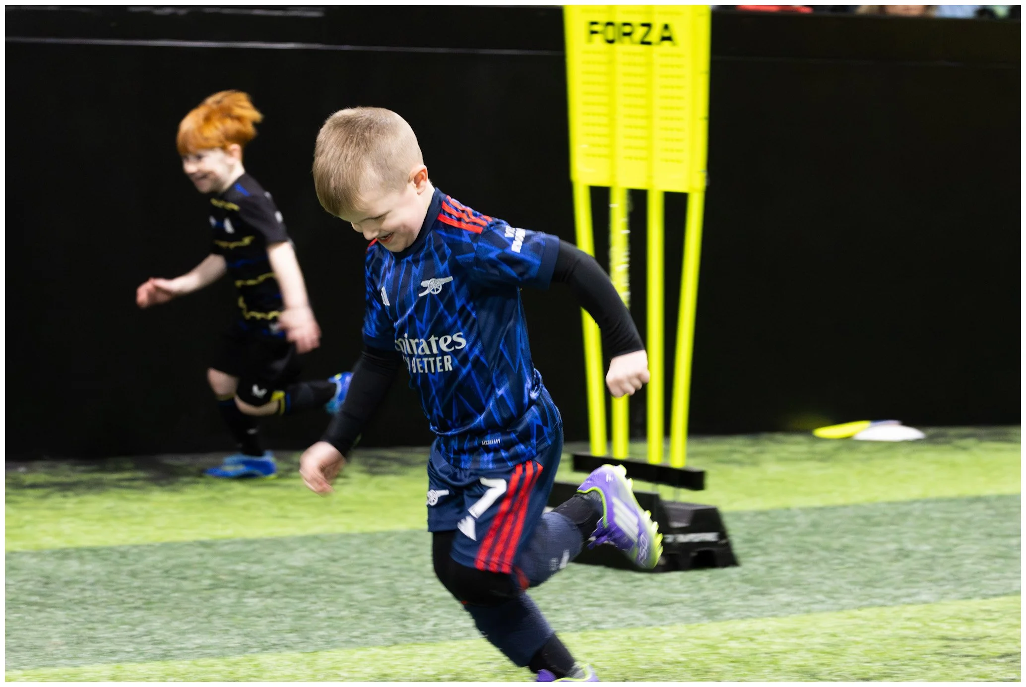 Two young boys playing indoor soccer, with one in a blue jersey and the other in a black and yellow jersey, on a green turf field with training equipment visible.