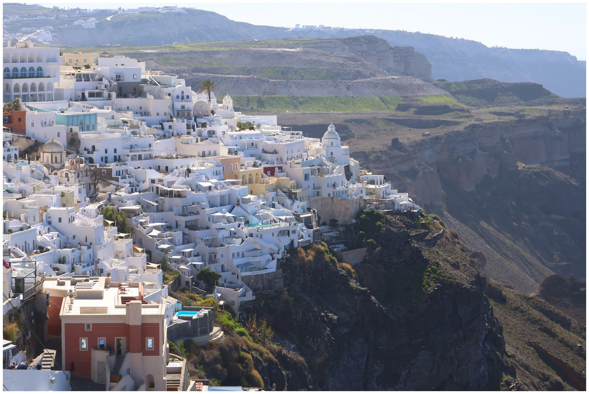 White buildings cascading down a hillside in Santorini, Greece, with a volcanic cliff face and distant green land in the background.