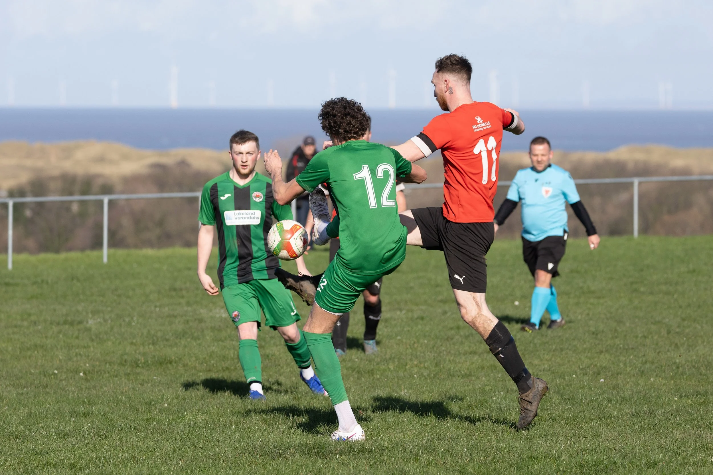 Two soccer players, one in a green uniform with number 12 and the other in a red uniform with number 11, contest the ball on a grassy field, with a referee observing in the background and a coastal landscape with wind turbines in the distance.