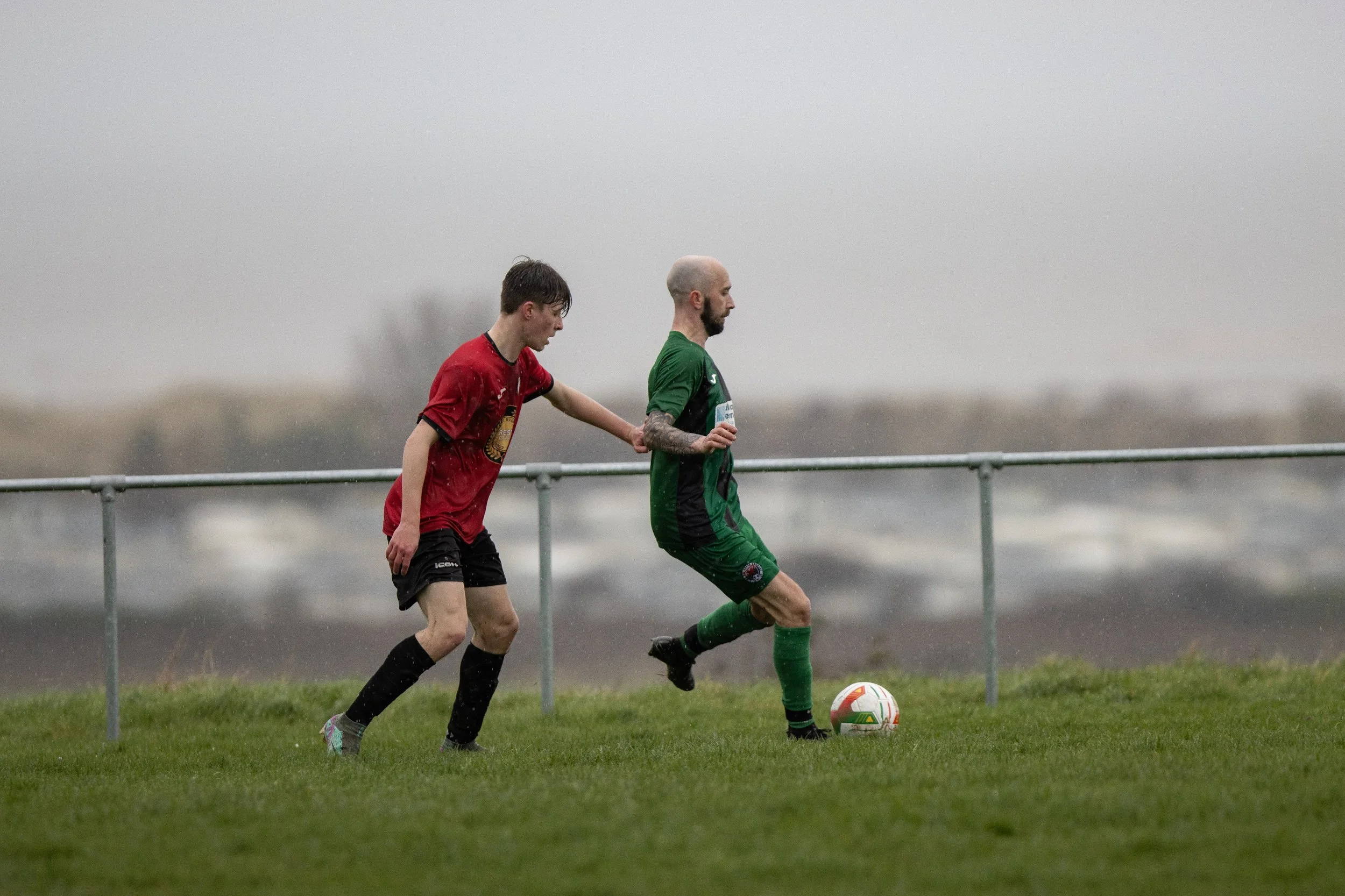 Two soccer players, one in a red jersey and the other in a green jersey, playing on a grassy field in rainy weather, with the player in green controlling the ball.