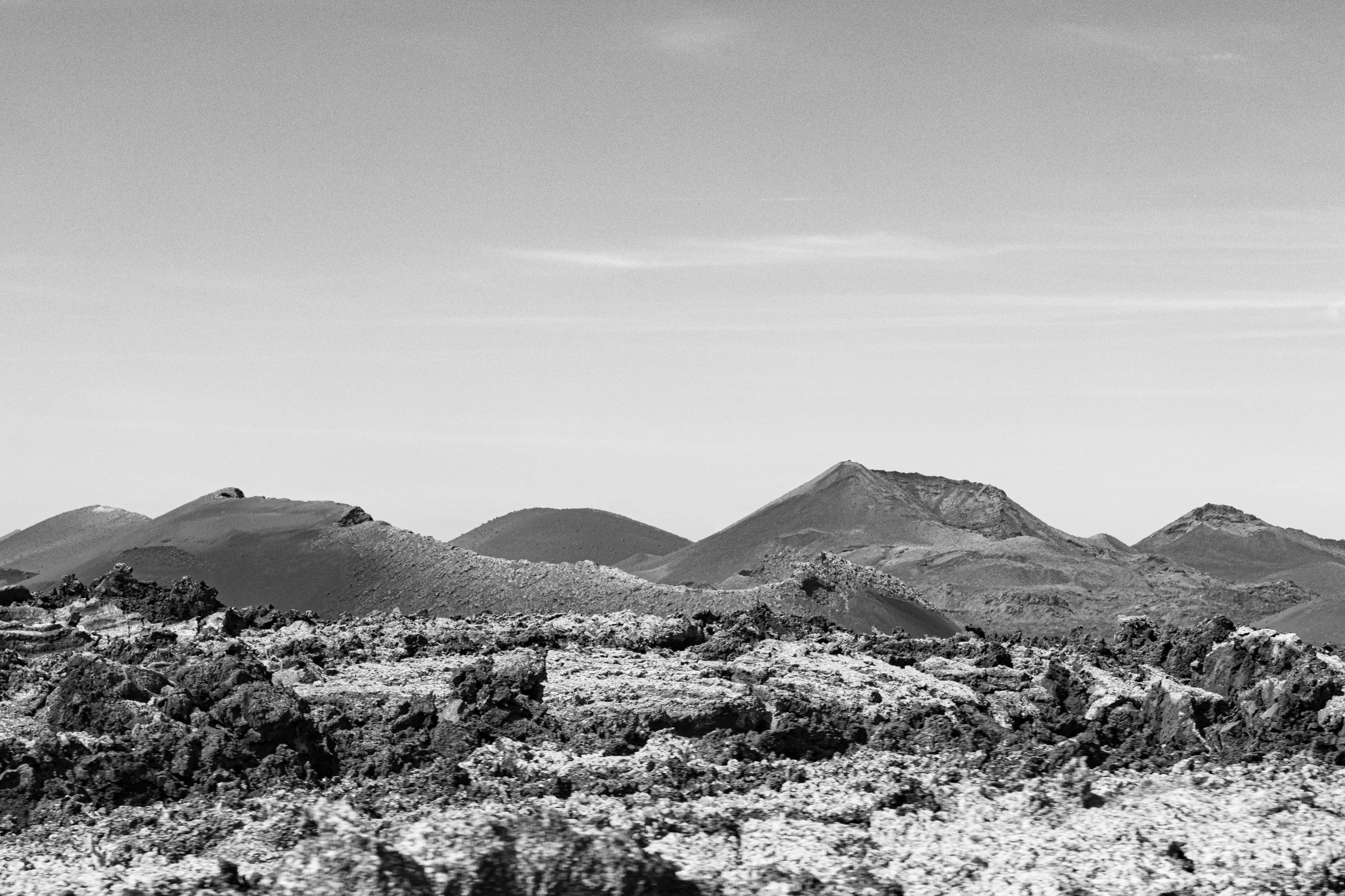 Black and white photograph of volcanic landscape with rugged, rocky foreground and multiple volcanic cones and mountains in the background under a clear sky.