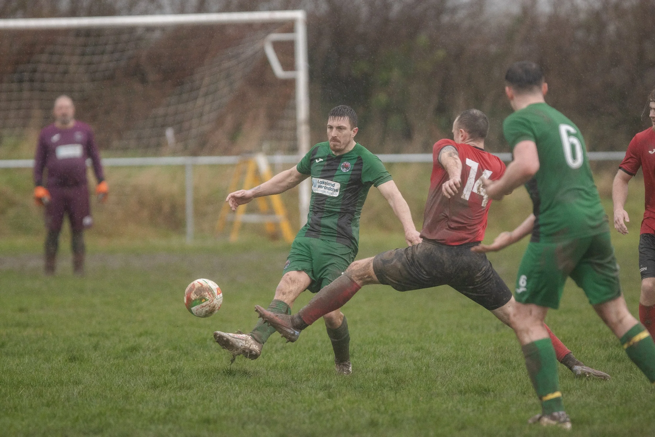Soccer game in rain, players in green and red jerseys competing for the ball, goalkeeper in purple in the background.