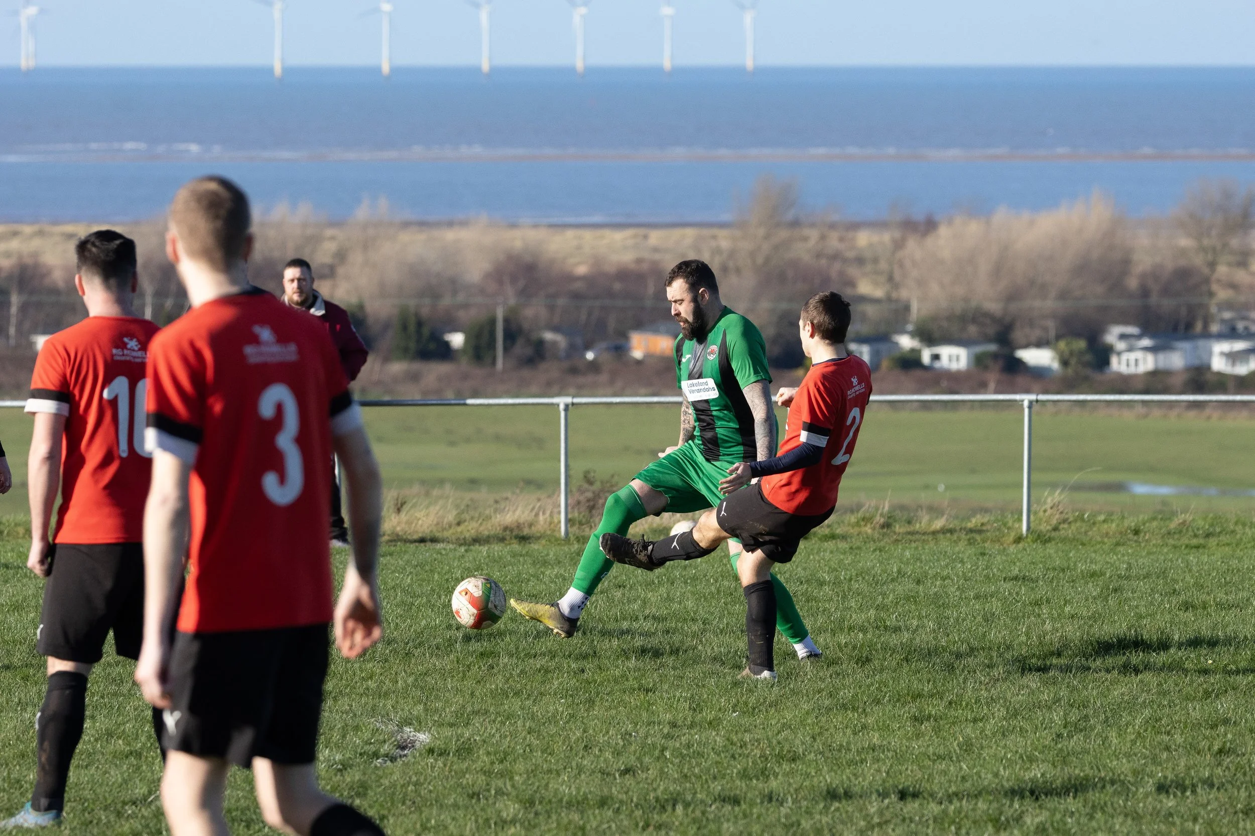 A soccer match taking place on a grassy field with a scenic background of water and wind turbines. Several players are engaged, with one in a green uniform and others in red and black uniforms.