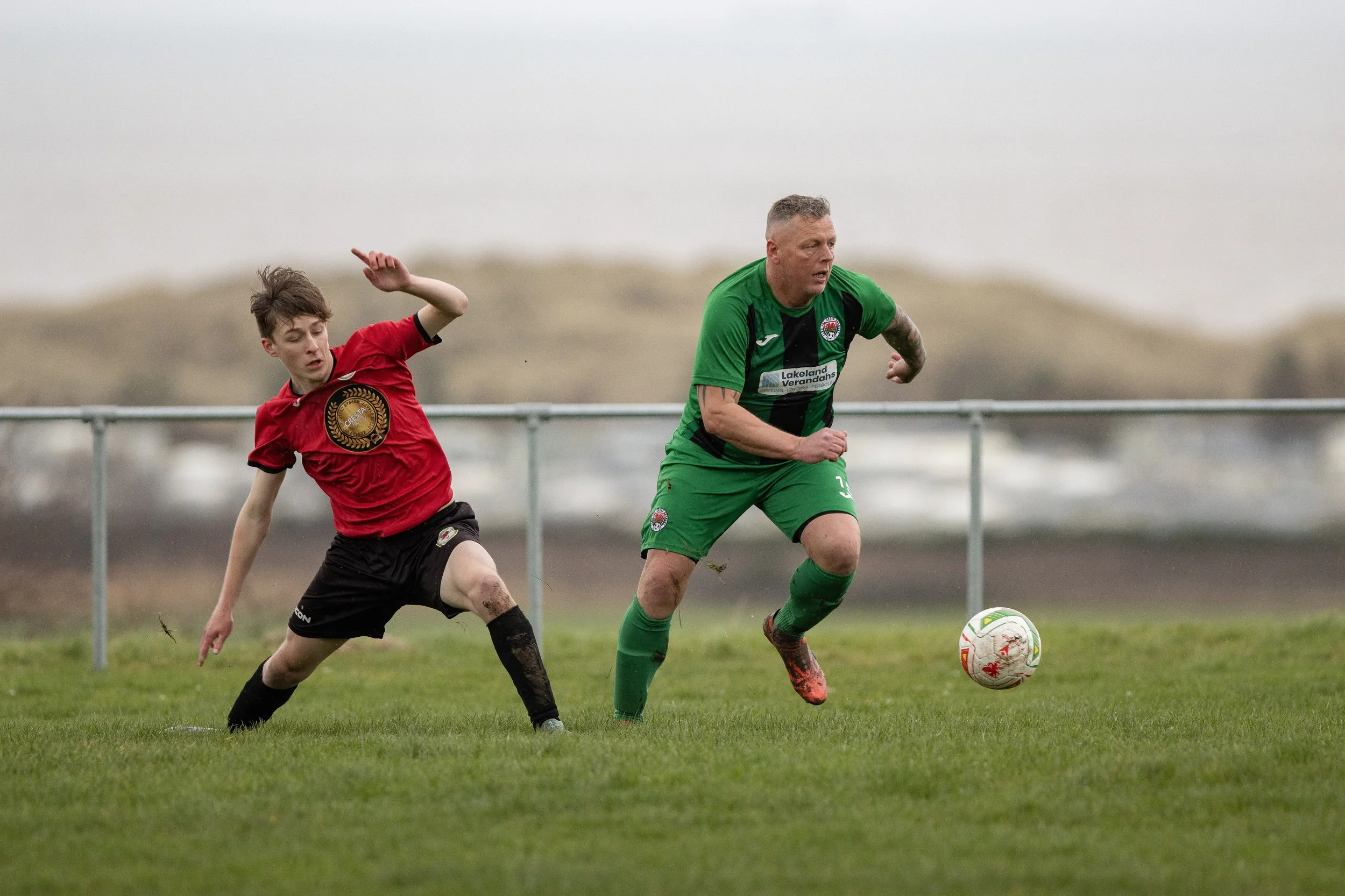 Two soccer players in action on a grassy field with a metal railing in the background. One player in a red jersey and black shorts is falling or diving, while the other player in a green jersey and shorts is running and about to kick the ball.