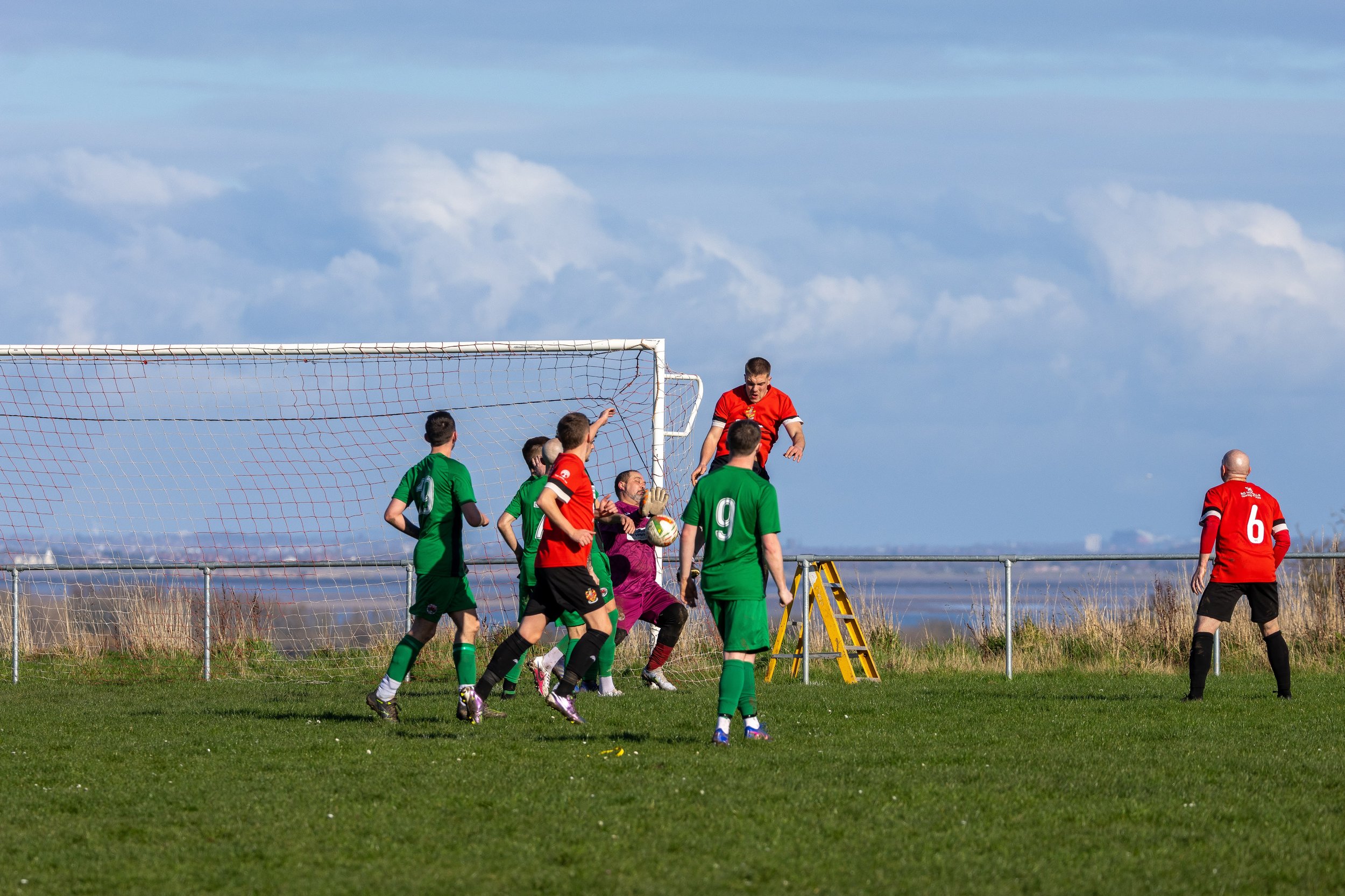 Soccer game showing players in red and green jerseys contesting near the goal with a goalkeeper in purple preparing for a save under a cloudy sky.