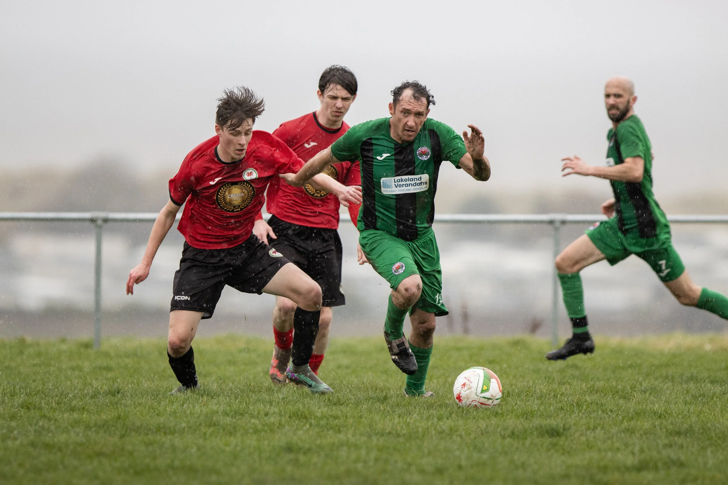 Soccer match with players in green and red jerseys on a rainy day on a grassy field.