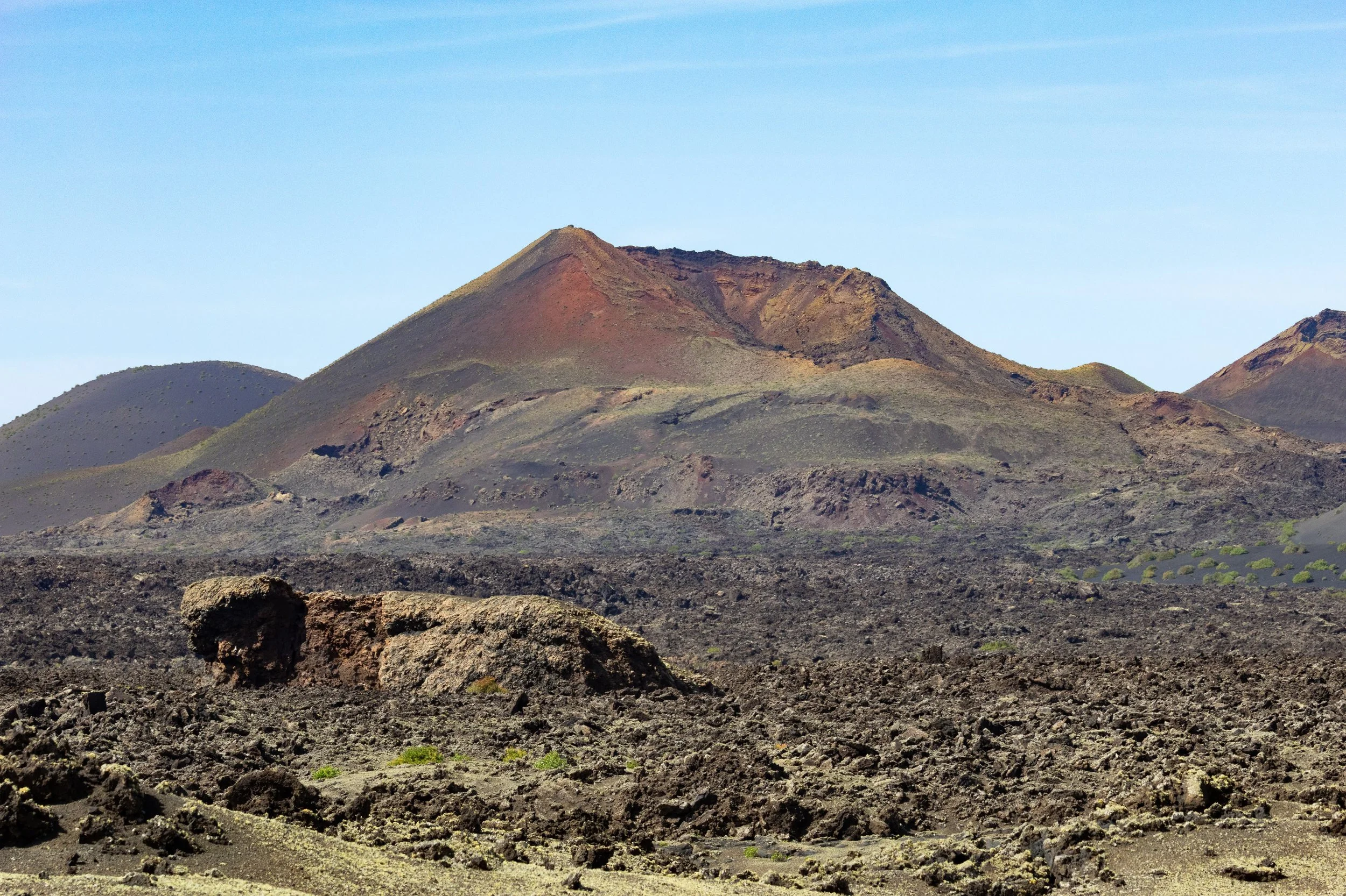 Landscape of volcanic terrain with dark, rocky ground and a large volcanic cone in the background under a clear blue sky.