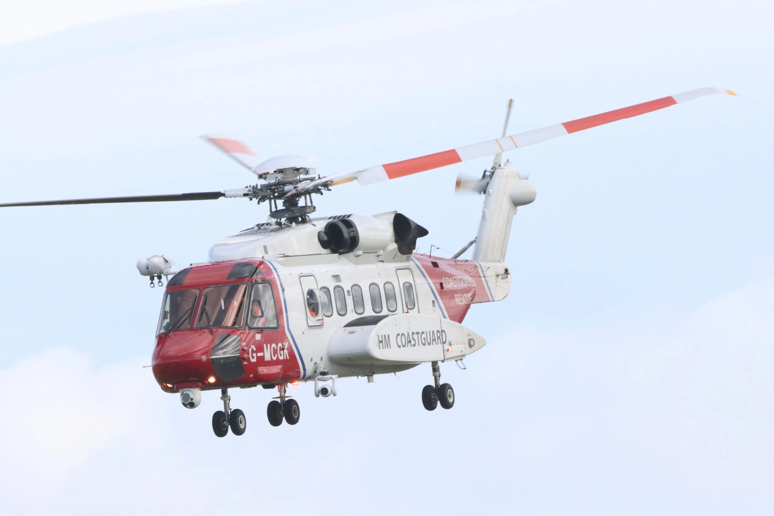 A rescue helicopter in flight with white and red colors, labeled 'HM COASTGUARD' and 'COASTGUARD RESCUE', flying against a cloudy sky.