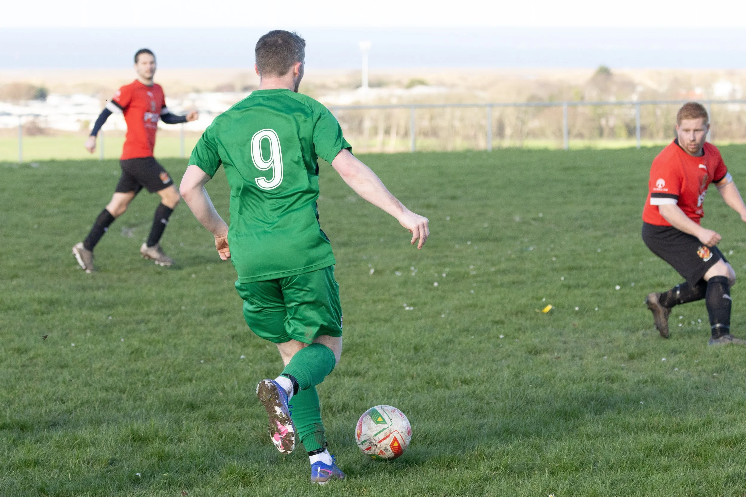 Soccer players on a field during a match. One player in a green uniform with the number 9 is about to kick the ball, while two players in red uniforms are nearby.