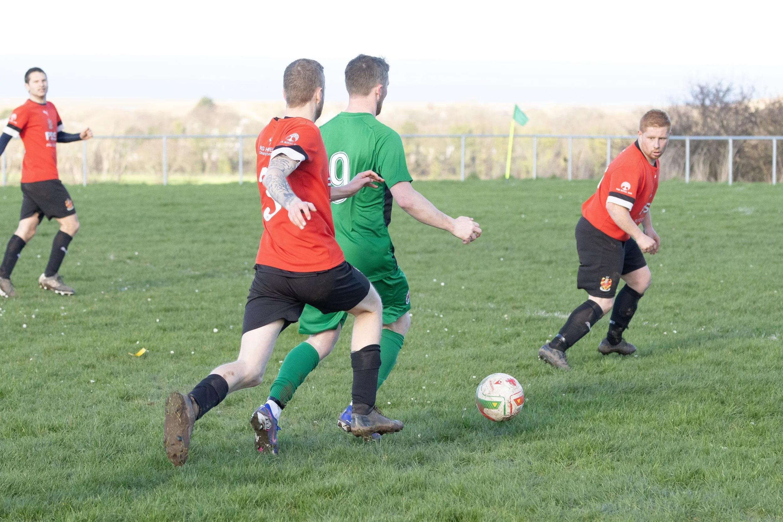 Soccer players in red and green jerseys chase a ball on a grassy field during a game.