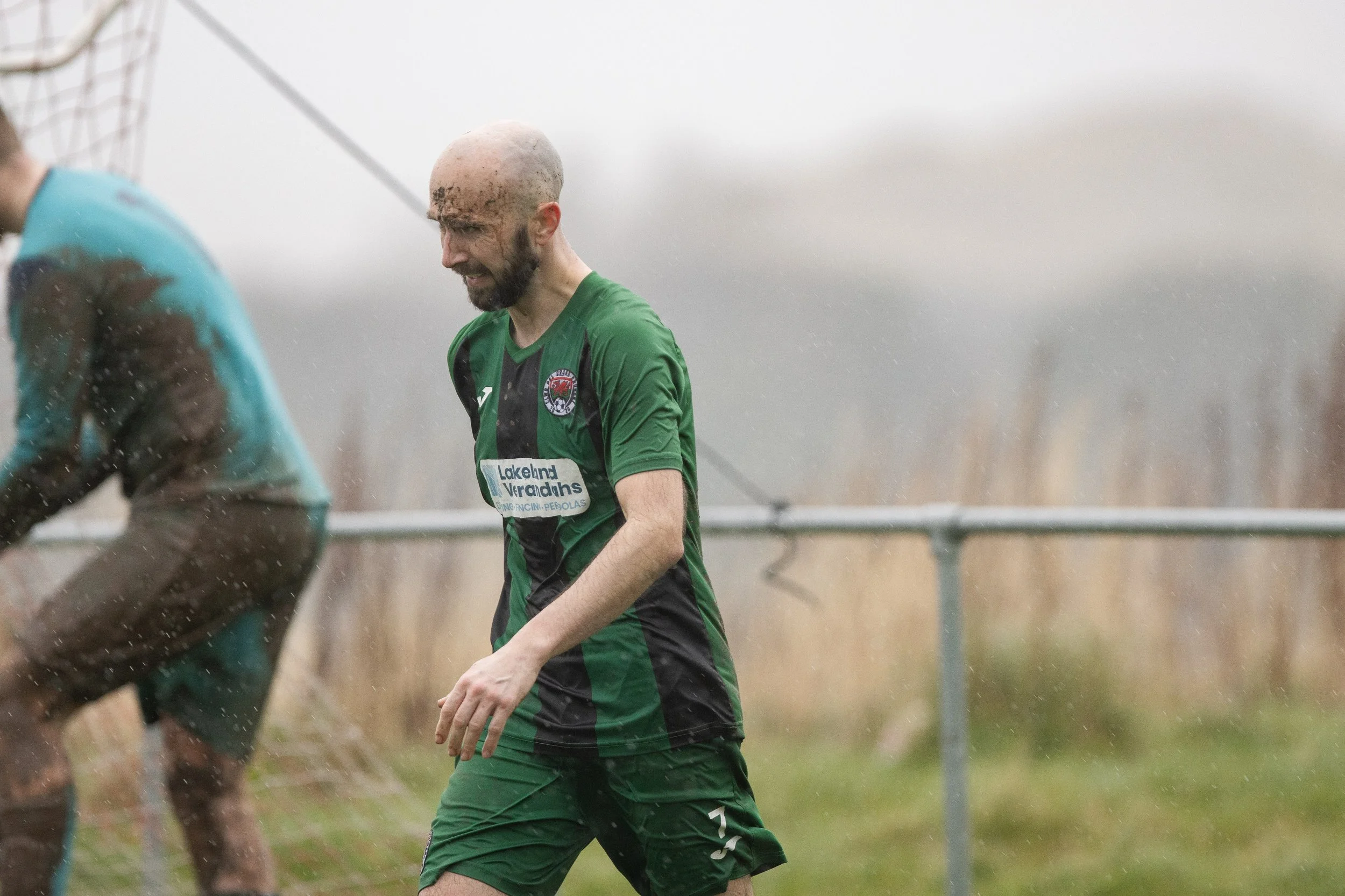 A soccer player wearing a green and black uniform with the number 7, standing on a wet field with a pained expression, sweat and mud on his face, and a bald head.