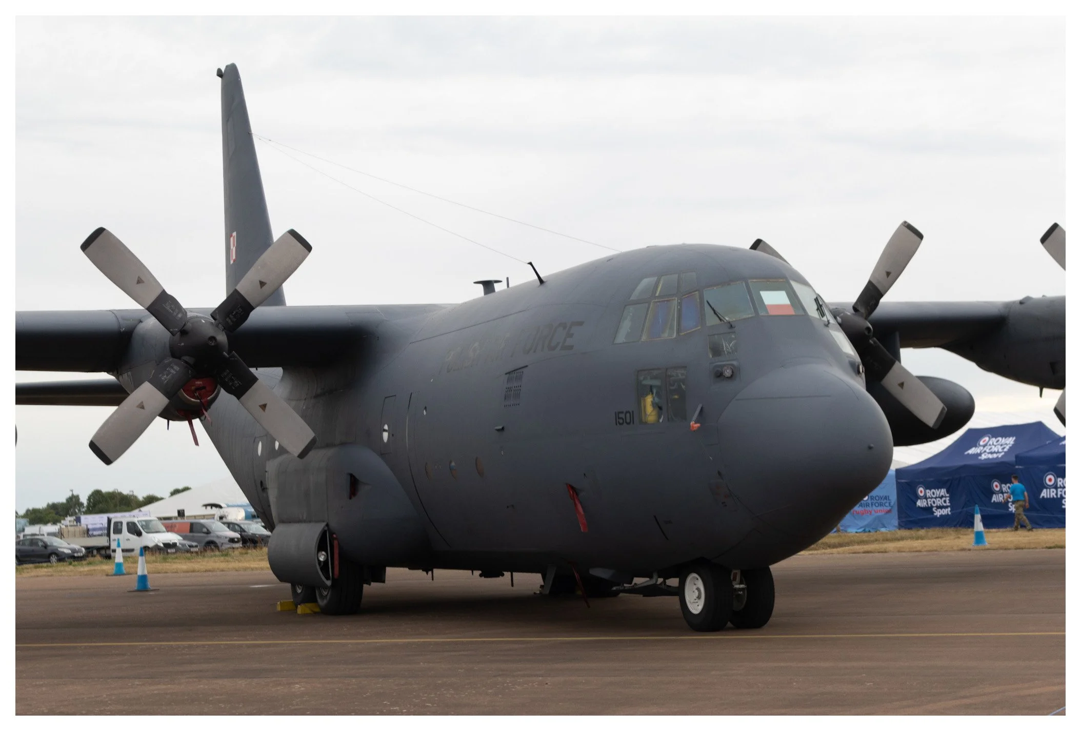 Gray military cargo aircraft on the tarmac with propellers and a cloudy sky above.