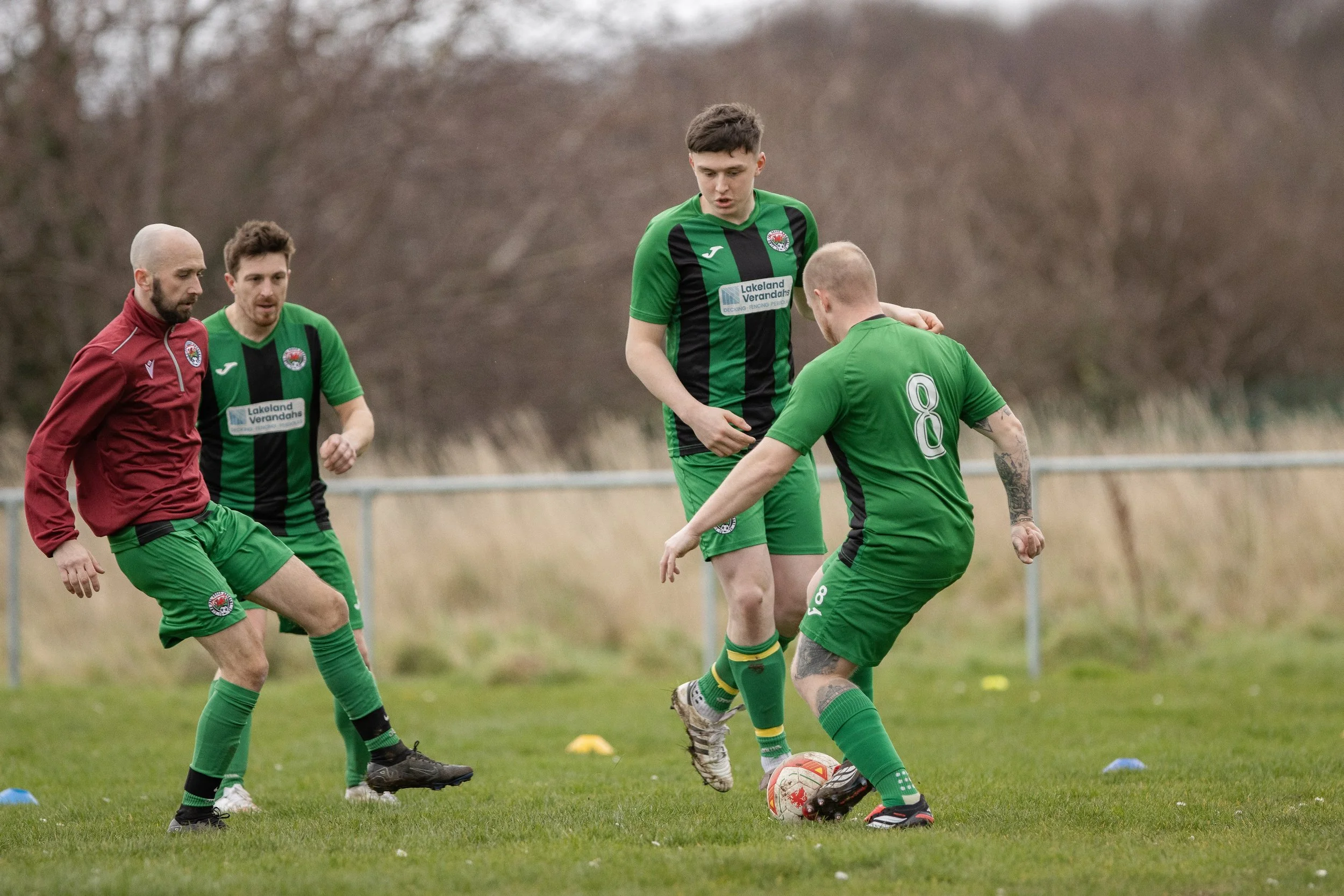 Four soccer players, three in green uniforms and one in a maroon top, playing on a grassy field during a practice session or game. One player with jersey number 8 is about to kick the ball while others observe.