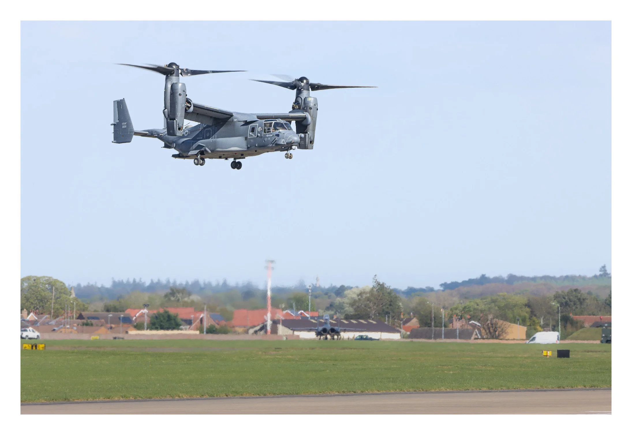 A gray military tiltrotor aircraft flying low over an airfield with a jet fighter on the runway and houses in the background.