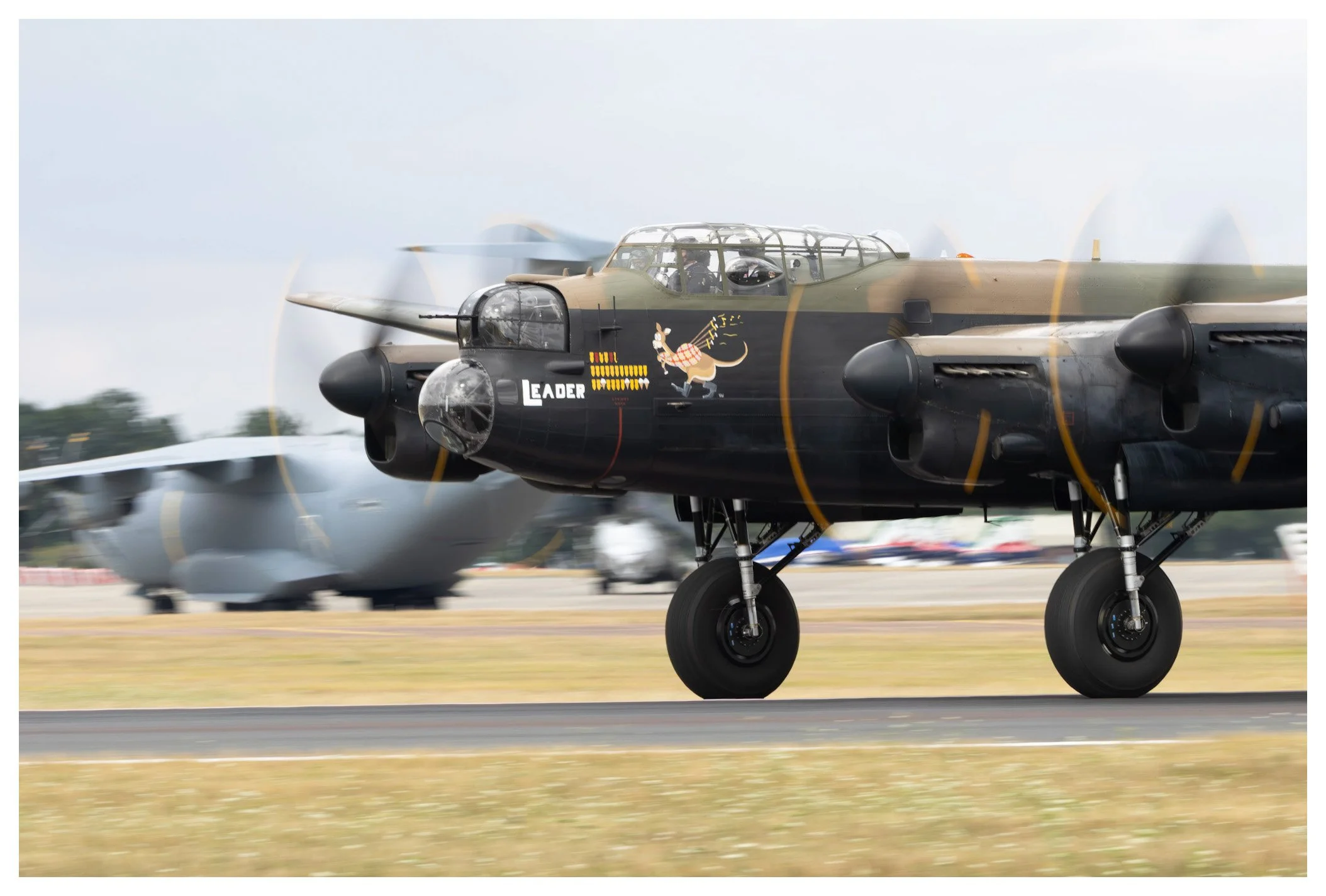 A military aircraft on the runway with a painted cartoon rabbit holding a paintbrush on its nose. The aircraft is black and gray with labeled markings, and other aircraft are blurred in the background.