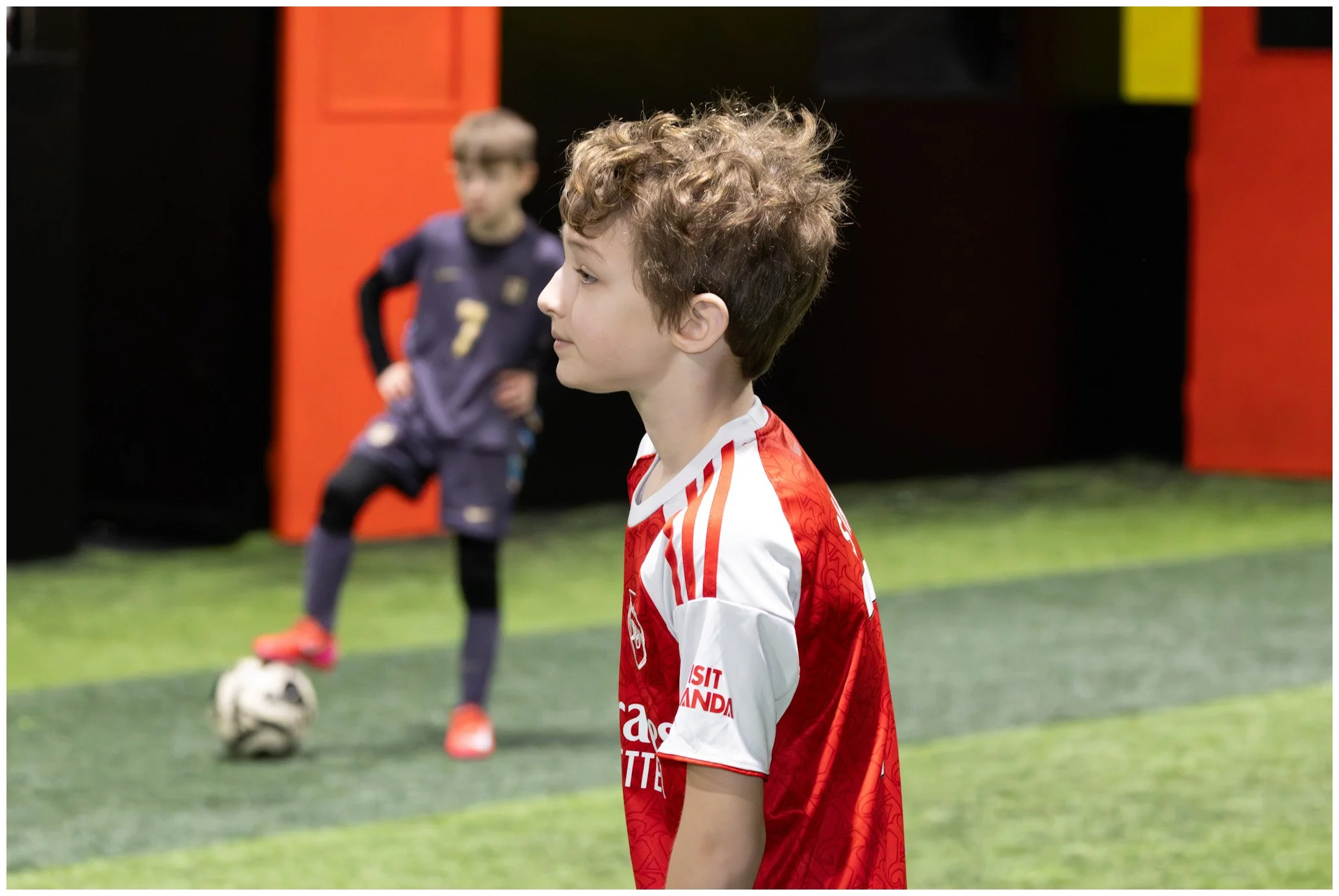 A young boy in a red and white soccer jersey stands on a soccer field, with a goalkeeper in black outfit in the background preparing to kick a soccer ball.