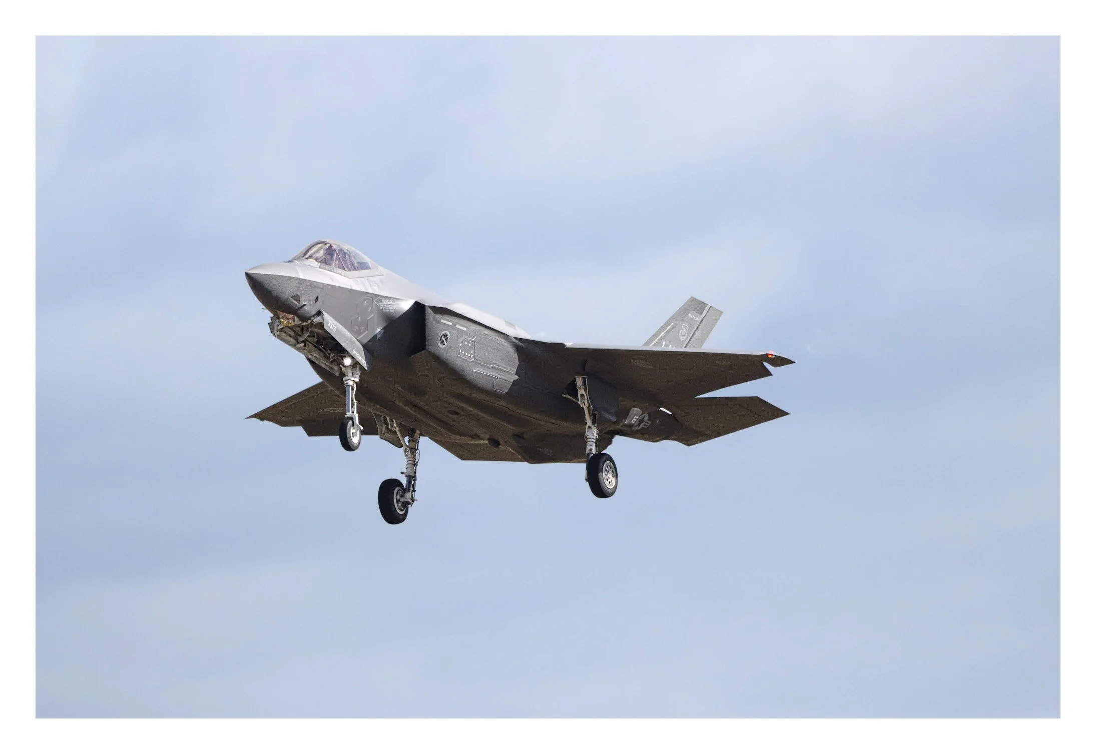 F-35 fighter jet in flight with landing gear extended against a cloudy sky.