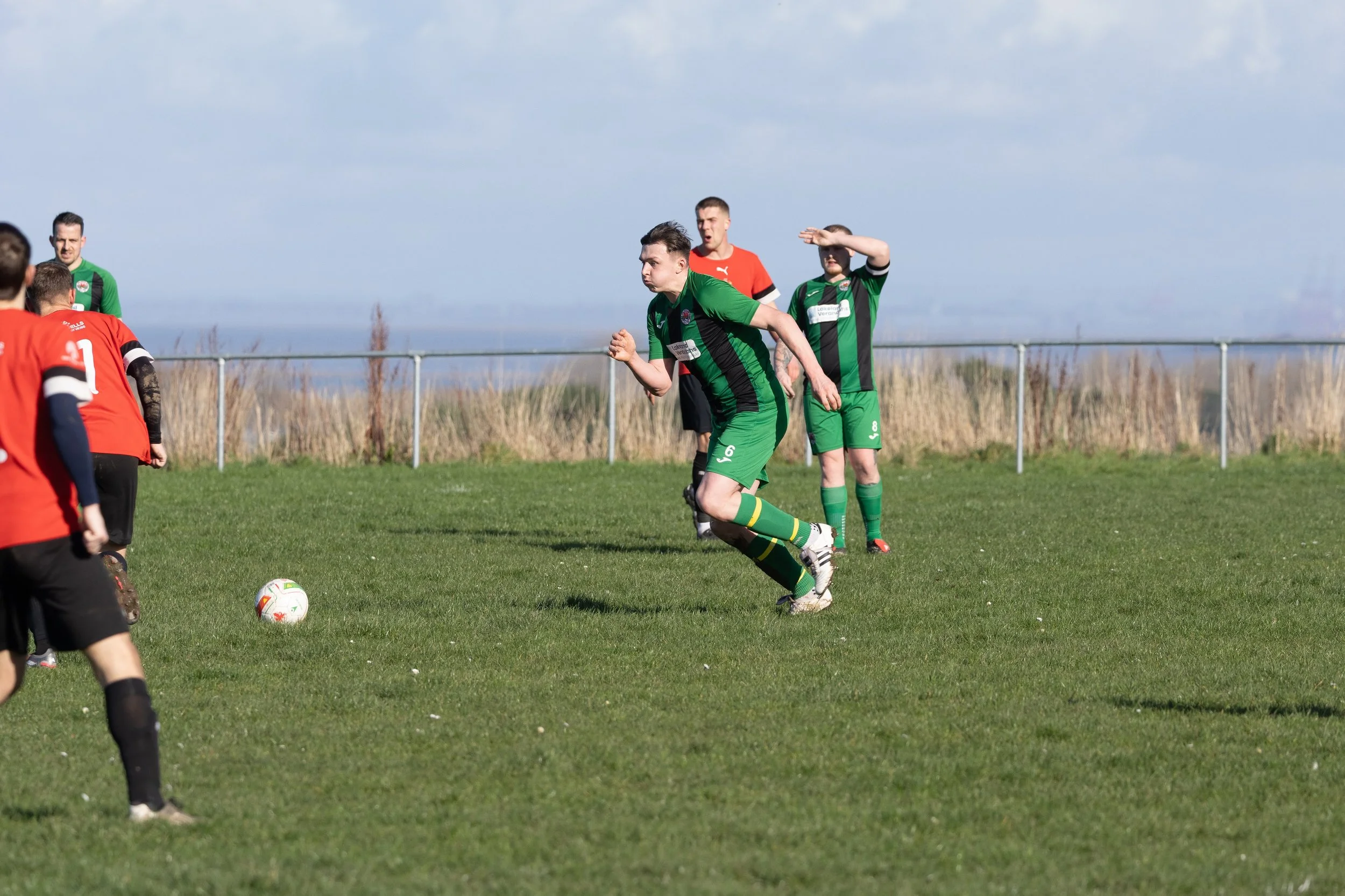 A soccer game with players in green and red uniforms on a grassy field, with a fence and open landscape in the background.