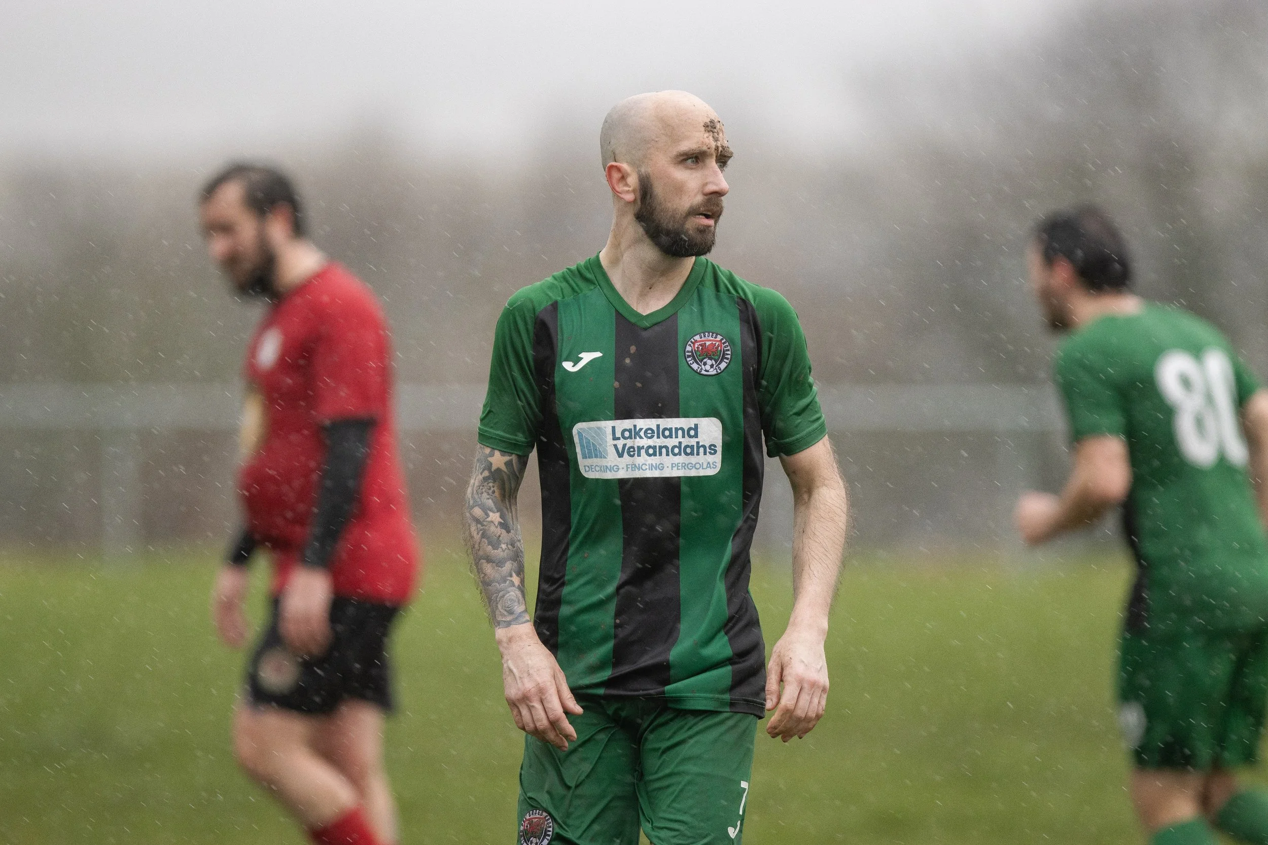Soccer players on the field during a game in rainy weather, with one player in the foreground wearing a green and black jersey, and two players in the background wearing red and green jerseys.