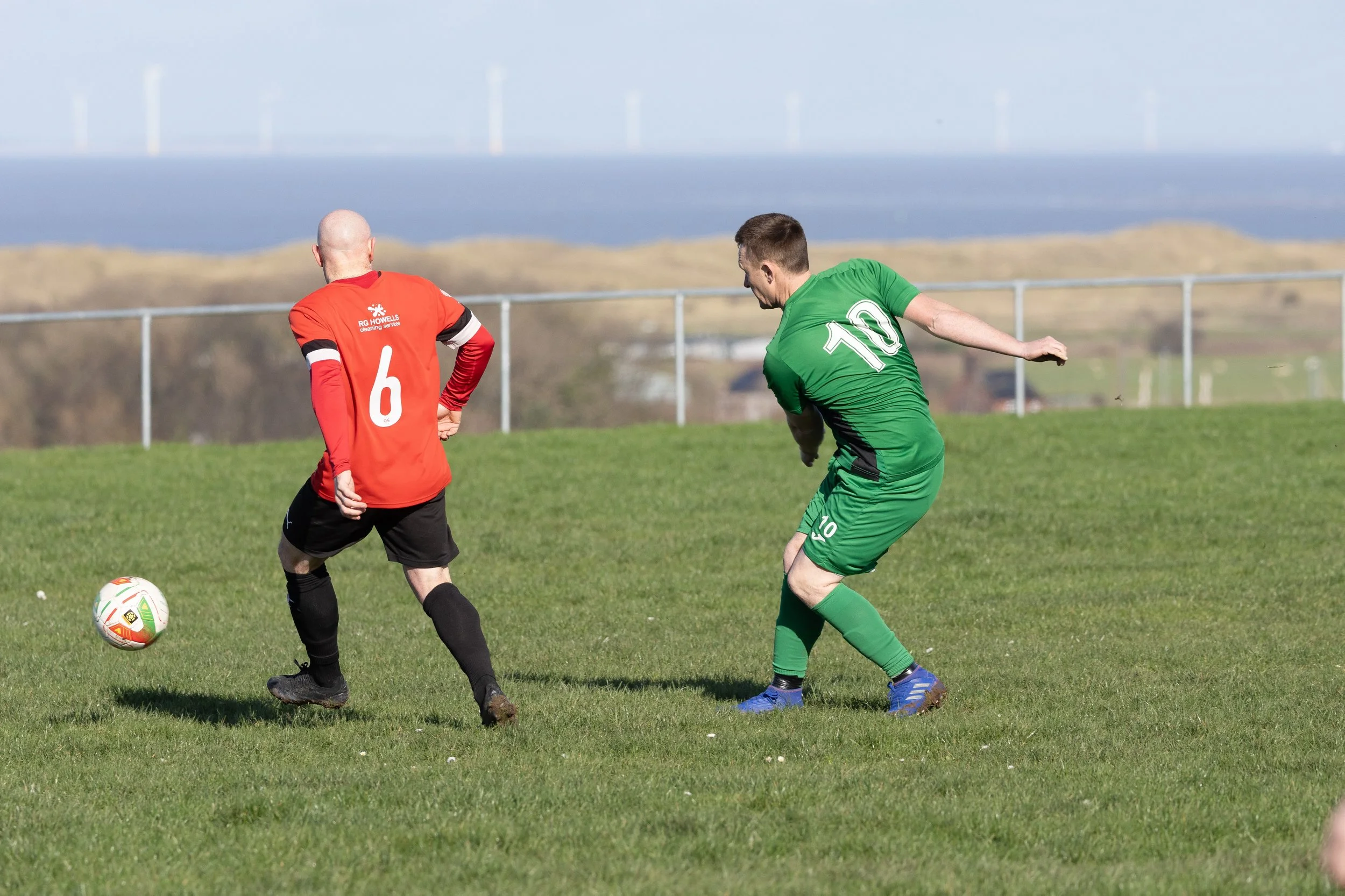 Two soccer players compete for the ball on the field, one in a red jersey with number 6, the other in a green jersey with number 10, during a match on a grassy field with a scenic landscape in the background.