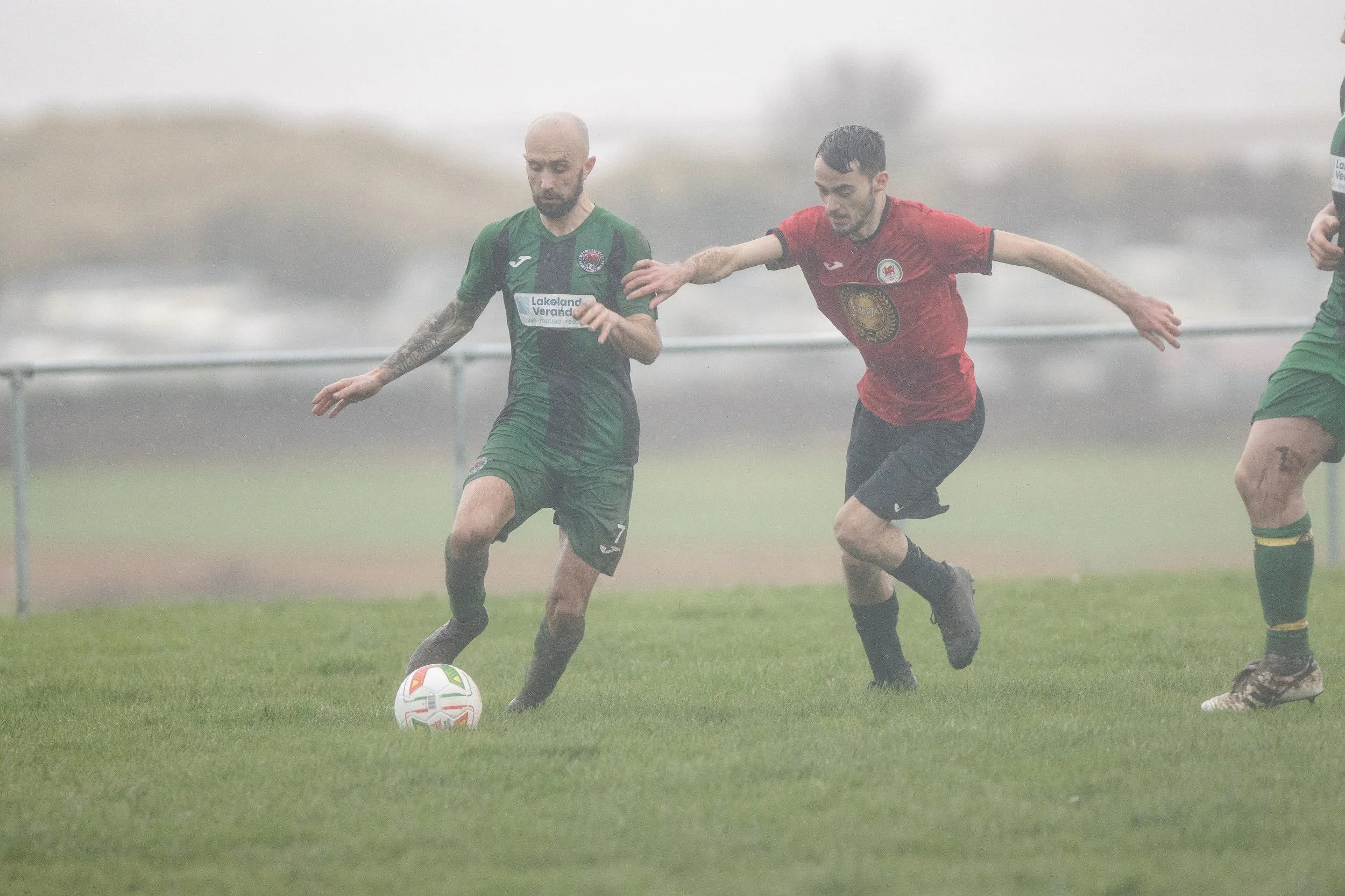 Two soccer players in green and red jerseys compete for the ball on a grassy field in rainy weather.