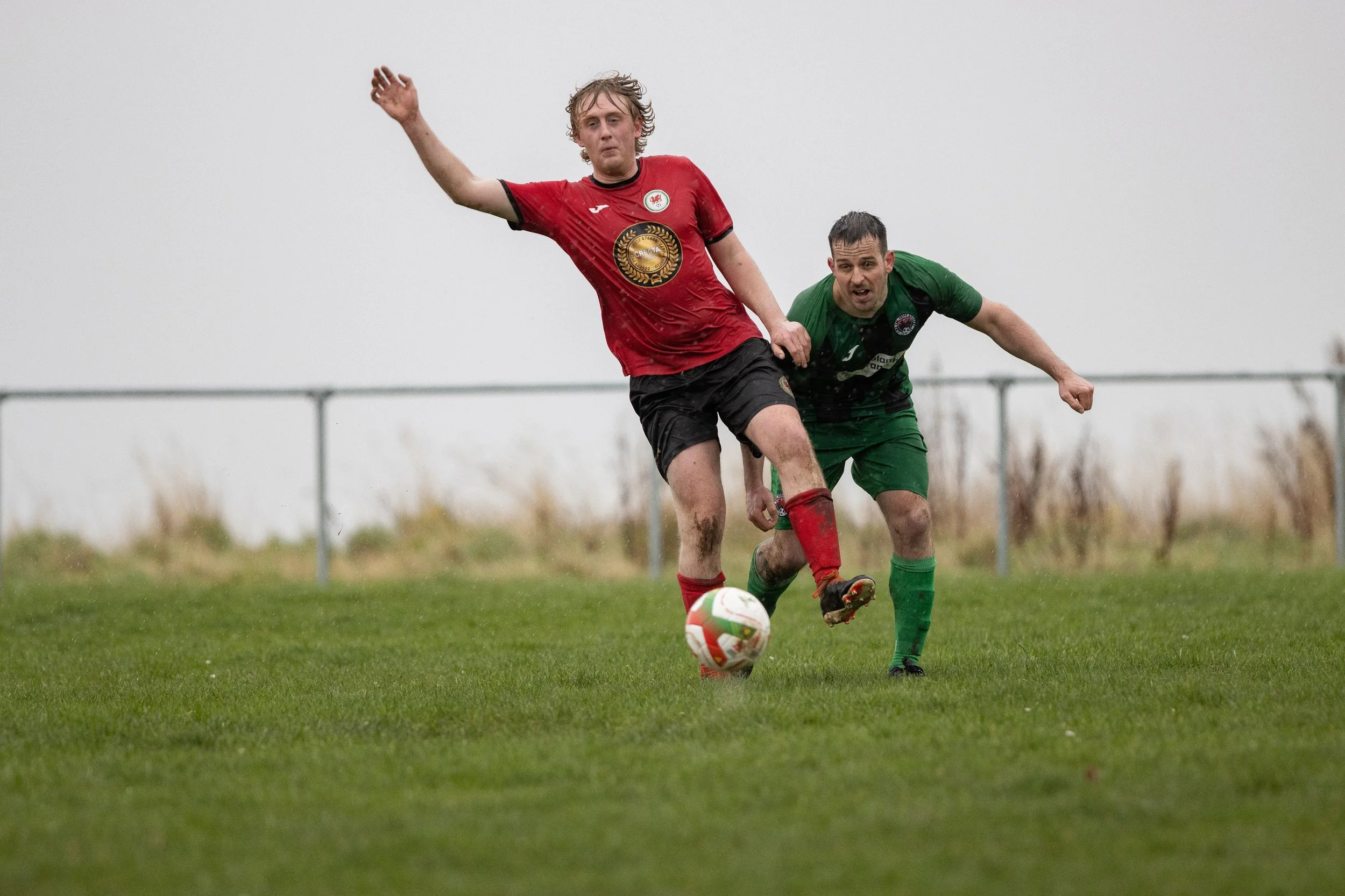 Two soccer players competing for the ball on a rainy field, one in a red jersey and black shorts, the other in a green jersey and shorts, with a fence in the background.