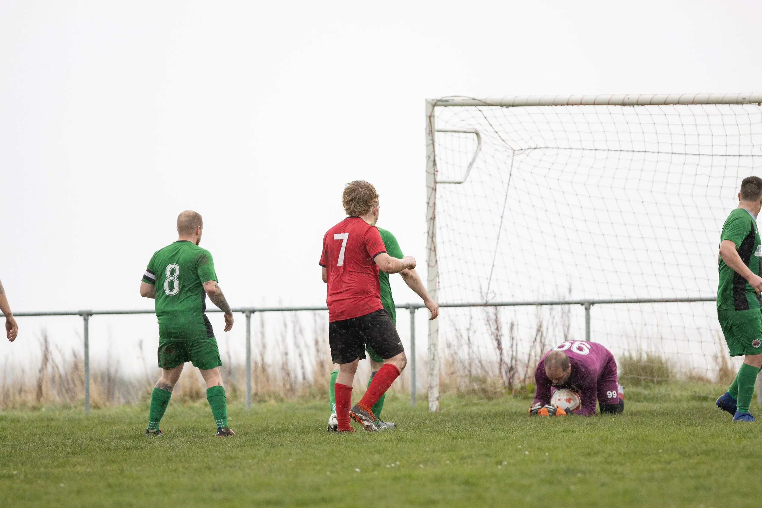 A soccer match with players in green and red jerseys, and a goalkeeper in purple, diving to save the ball near the goal on a grassy field.