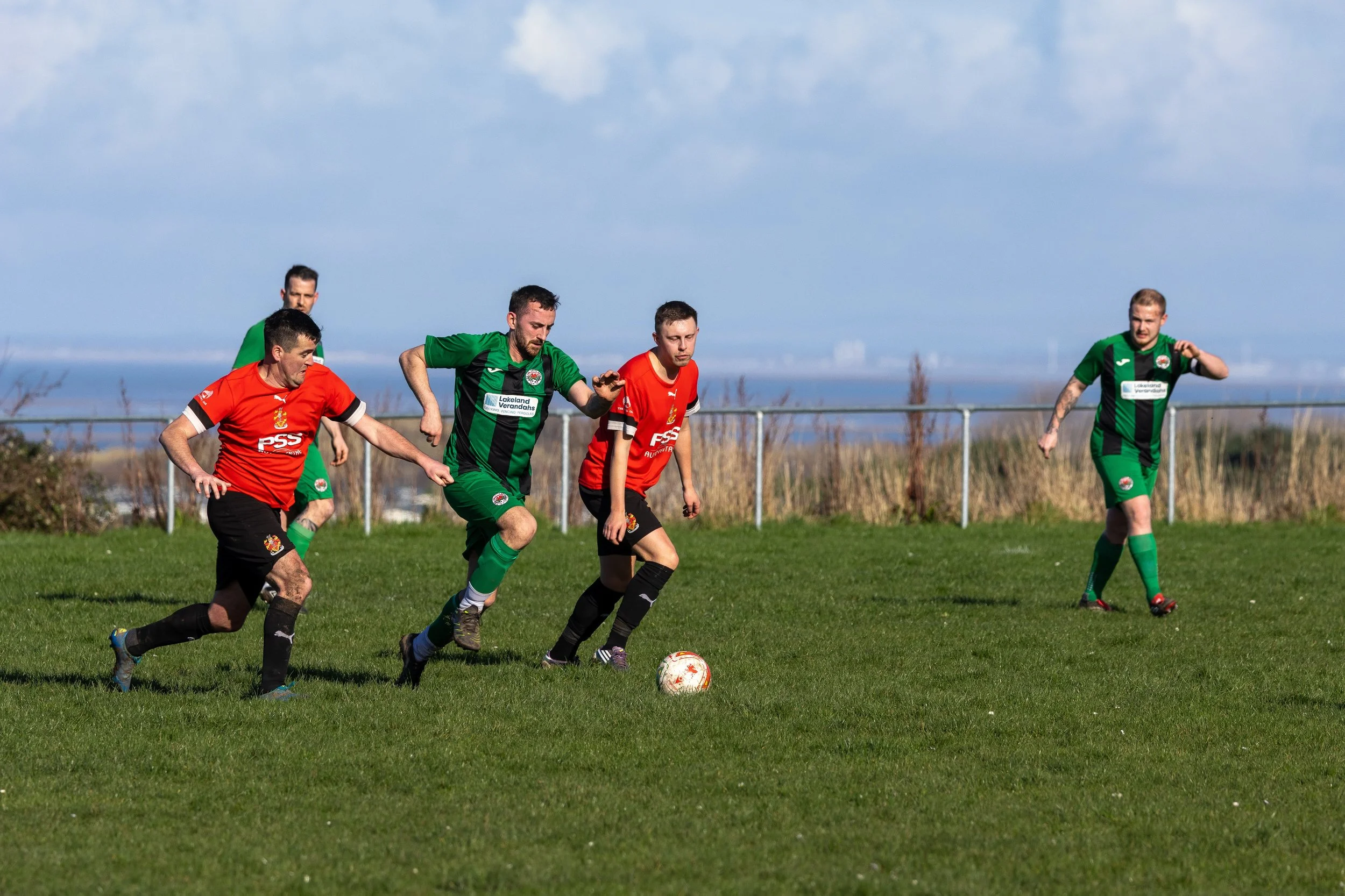 Soccer players competing for ball on field, with two players in red jerseys and three in green jerseys.