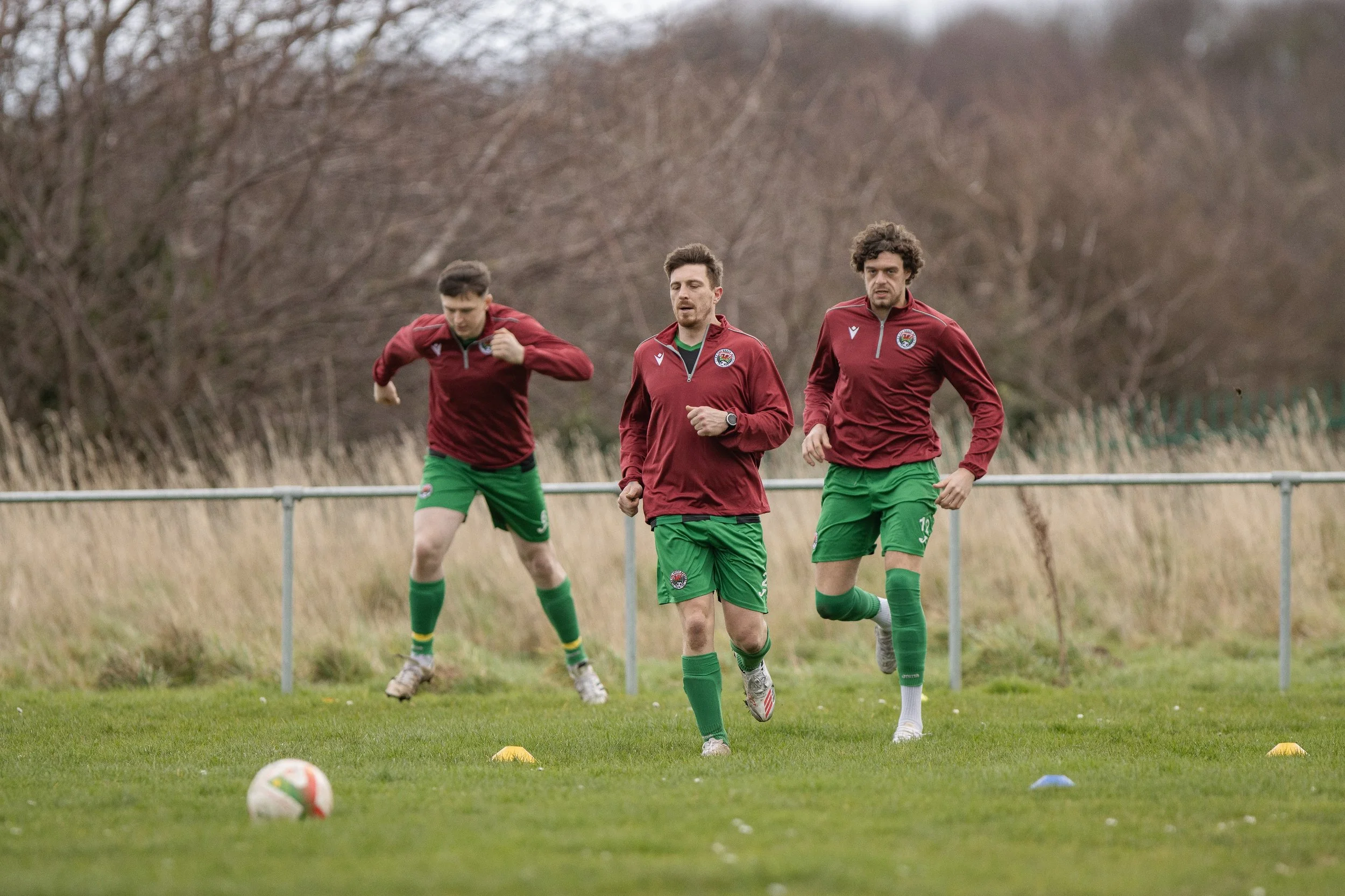 Three male soccer players jogging on the field during training, wearing maroon jackets and green shorts, with a soccer ball in the foreground and training cones on the grass.