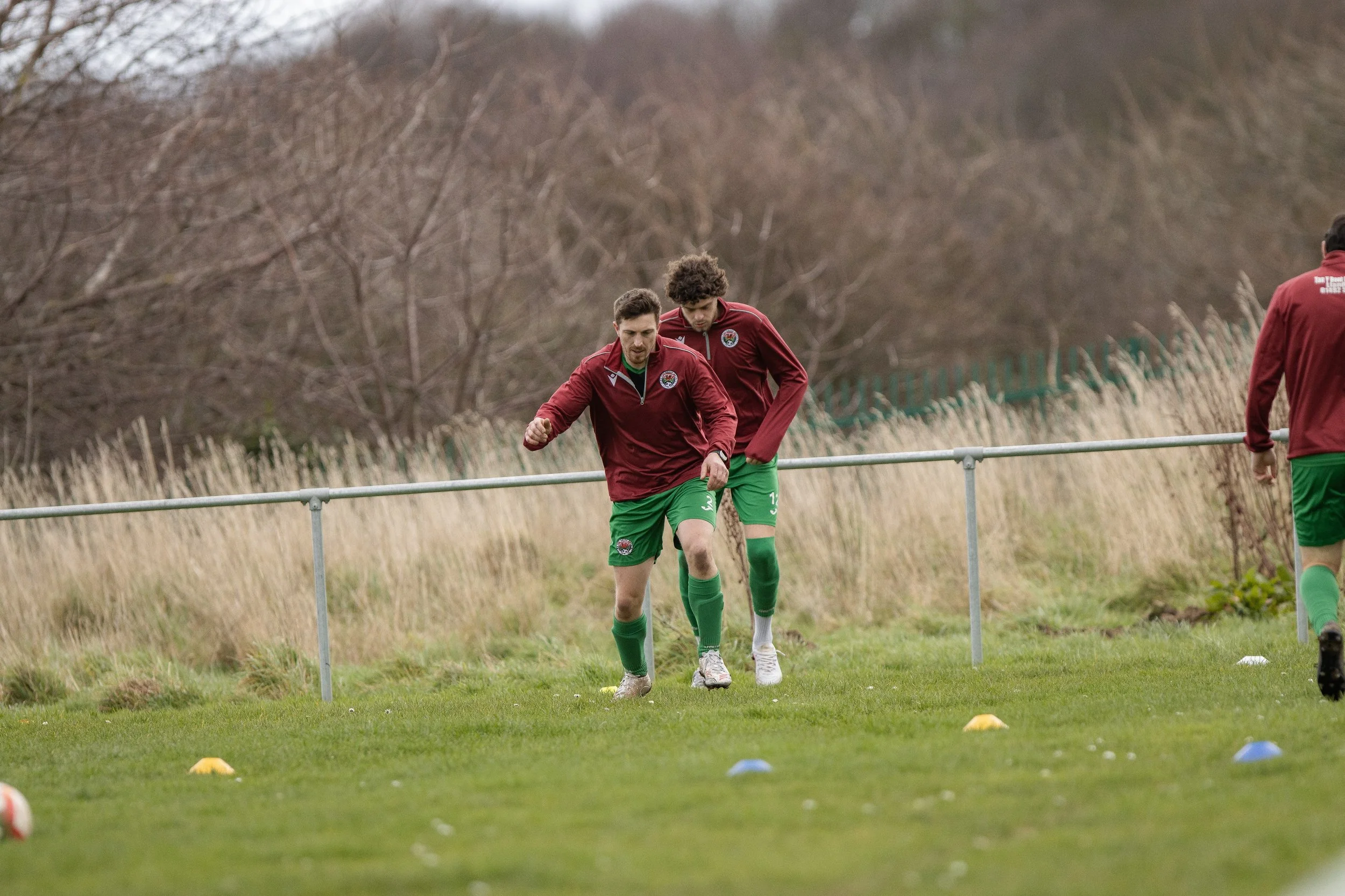 Two soccer players in maroon jackets and green shorts jogging on a grassy field during a training session with cones laid out on the ground.