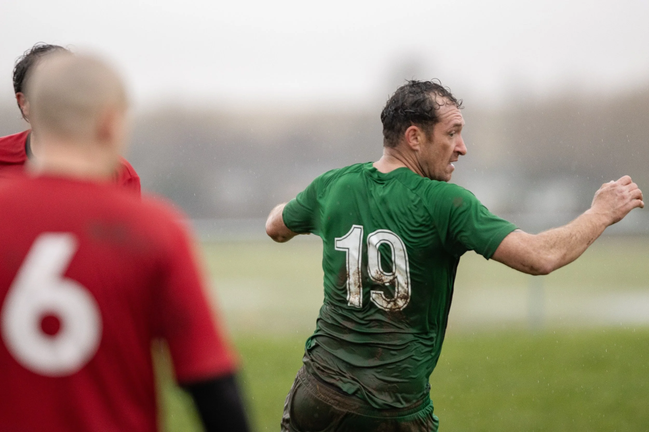 A soccer player wearing a green jersey with the number 19 on the back is running on the field in a rainy match. Two players in red jerseys are visible, one in the foreground and one in the background.