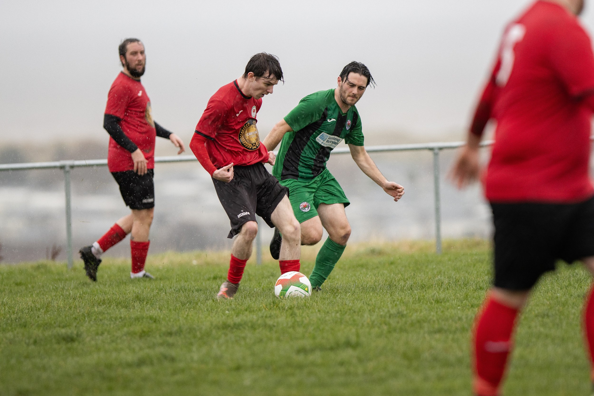 Two soccer players contest for the ball on a rainy field, with two additional players visible in the background.