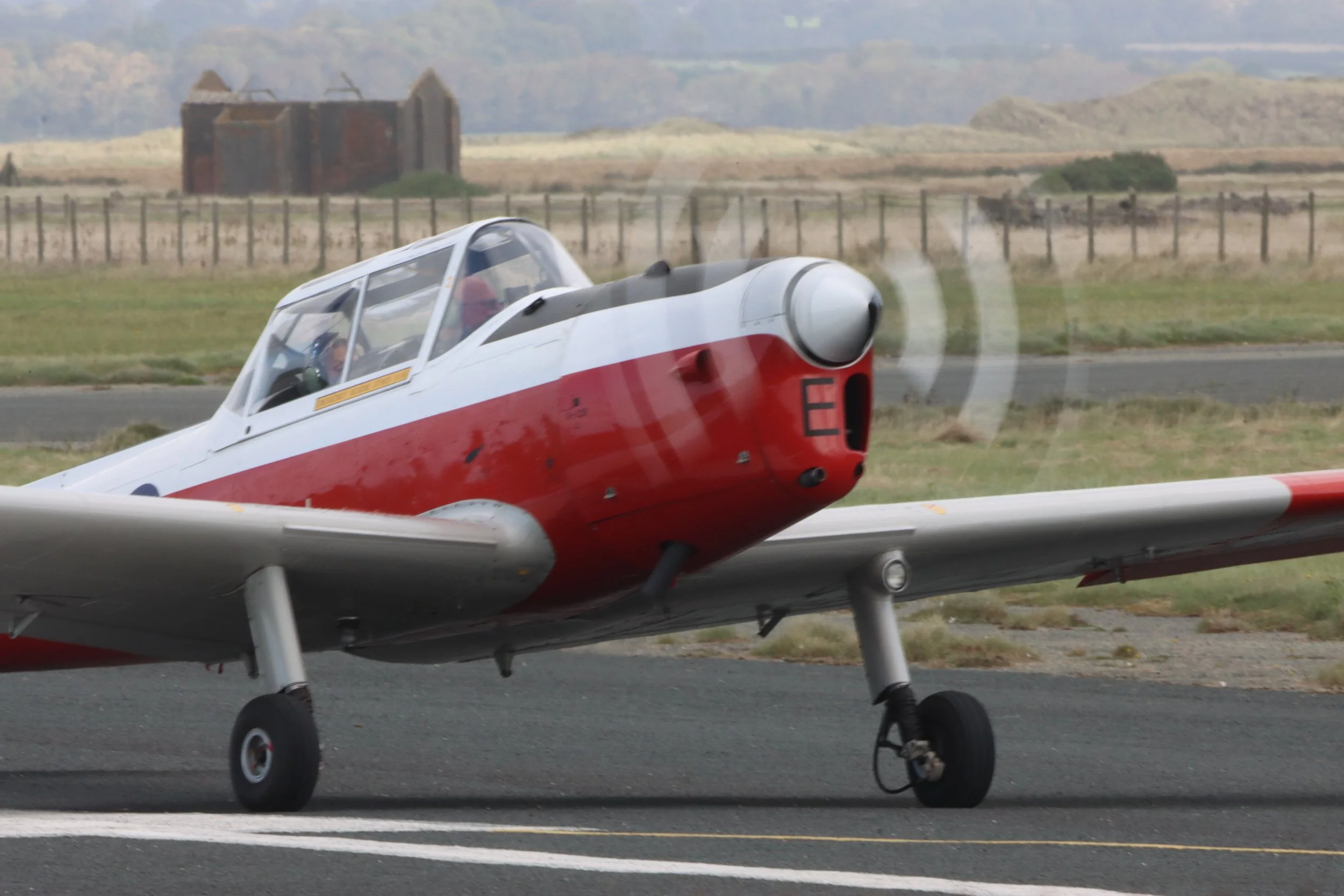 A vintage propeller airplane on a runway, with a red and white body, in a rural area with a fence and a distant building in the background.