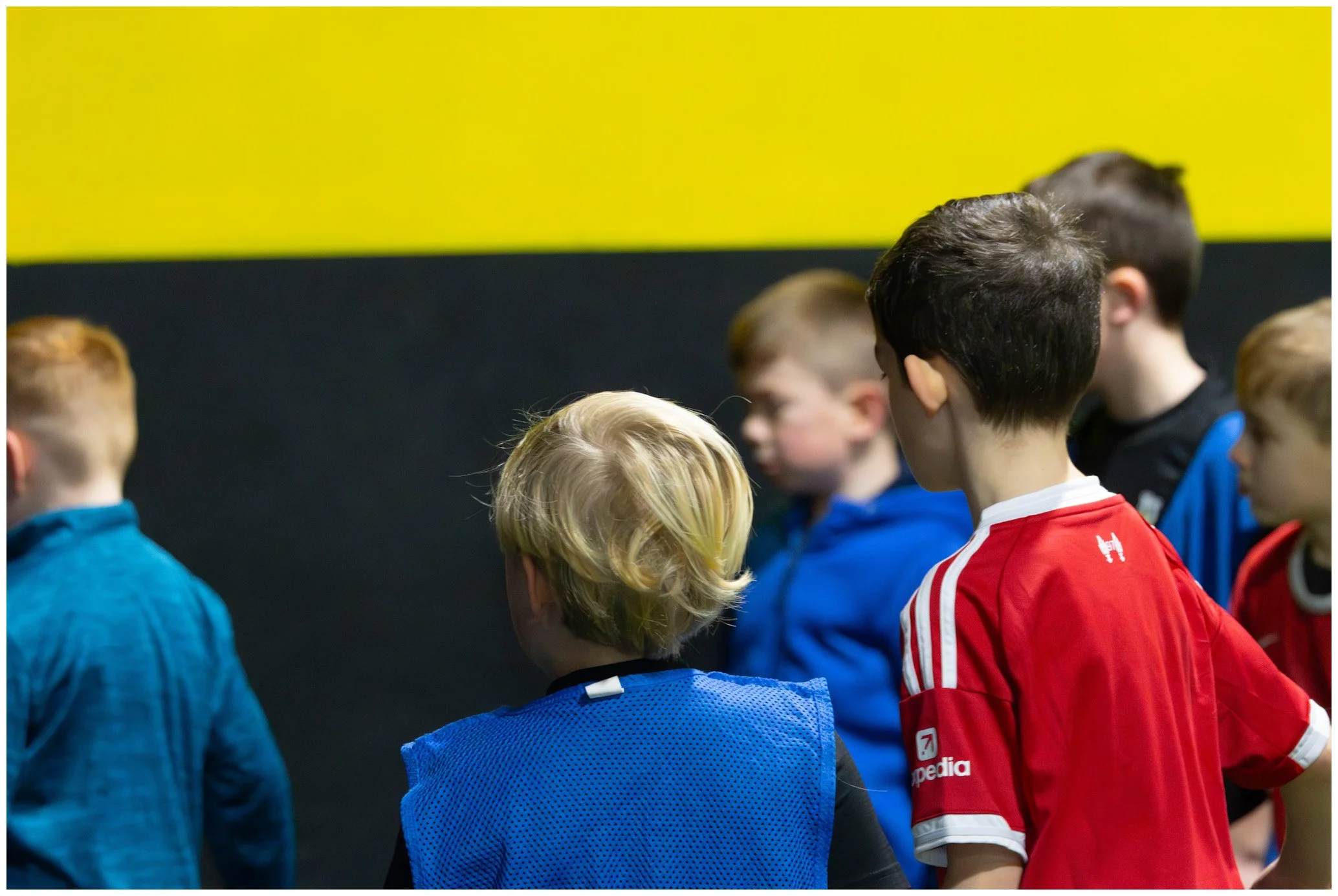 Group of young boys standing together, some facing to the side or back, wearing sports jerseys in red, blue, and black, indoors with a yellow and black wall in the background.