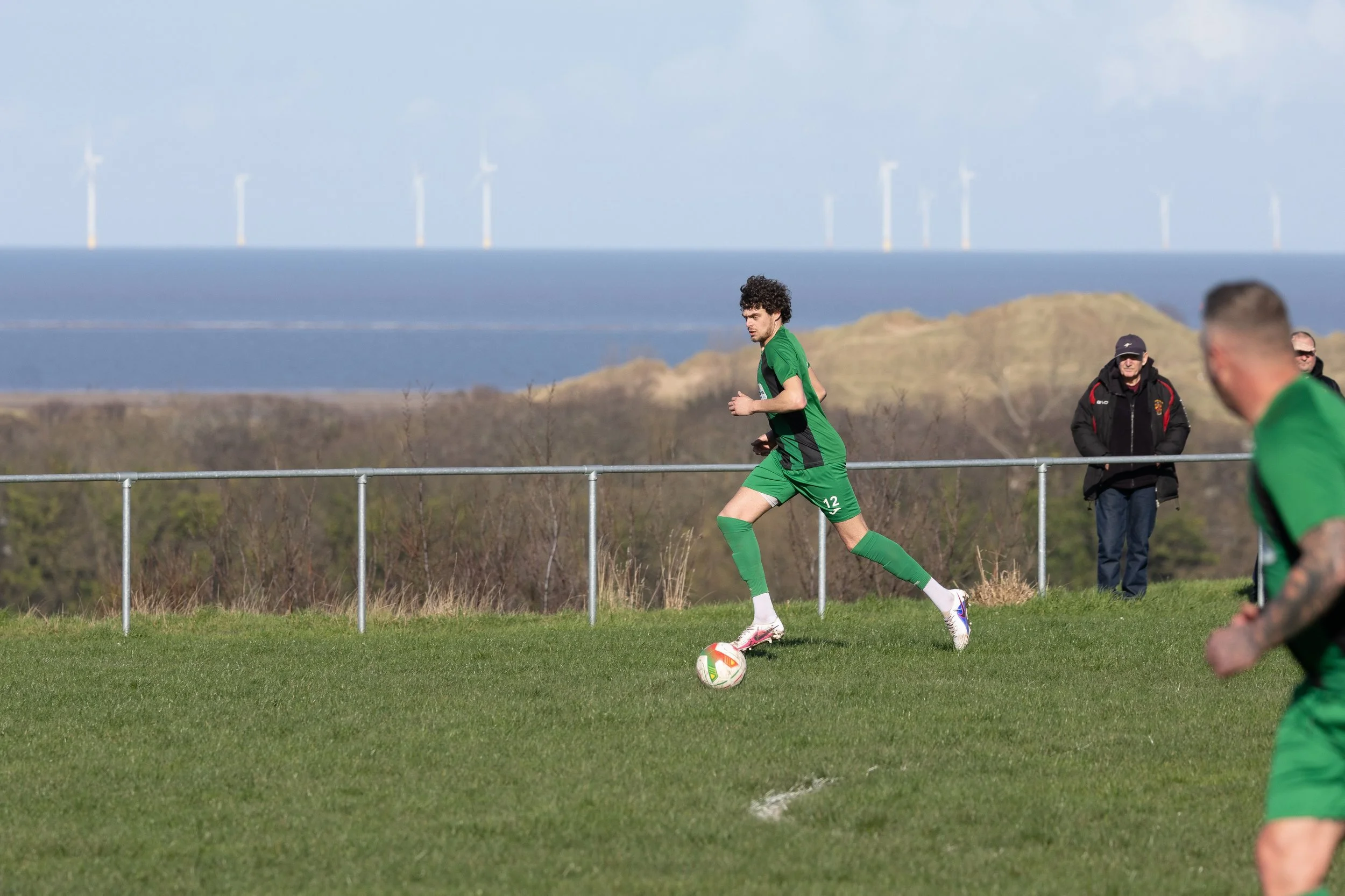 Soccer players in green jerseys playing on a grass field with a coastal landscape and wind turbines in the background.