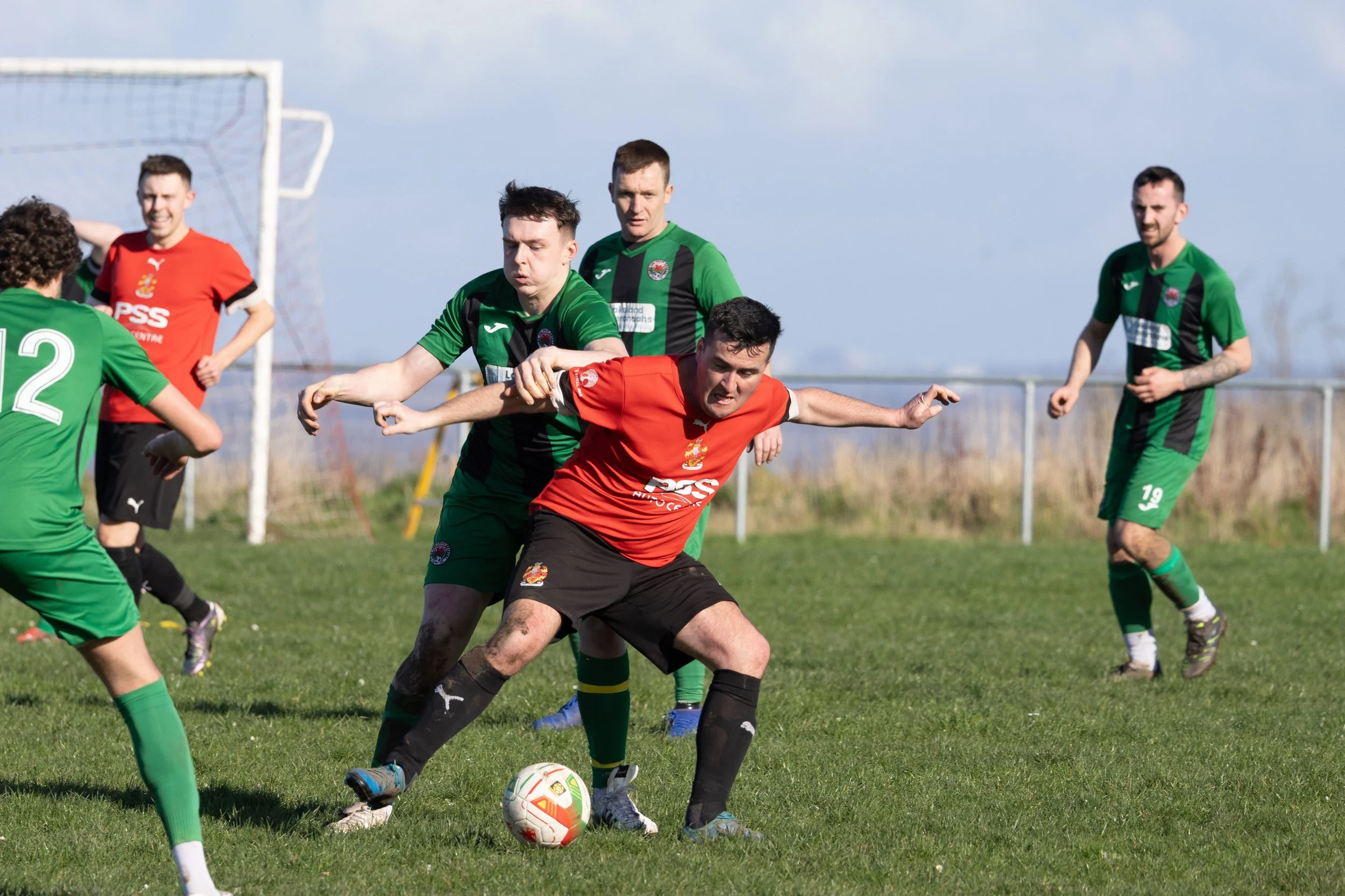 Soccer players competing for the ball on a grassy field, with a goalpost in the background, during a match on a sunny day.