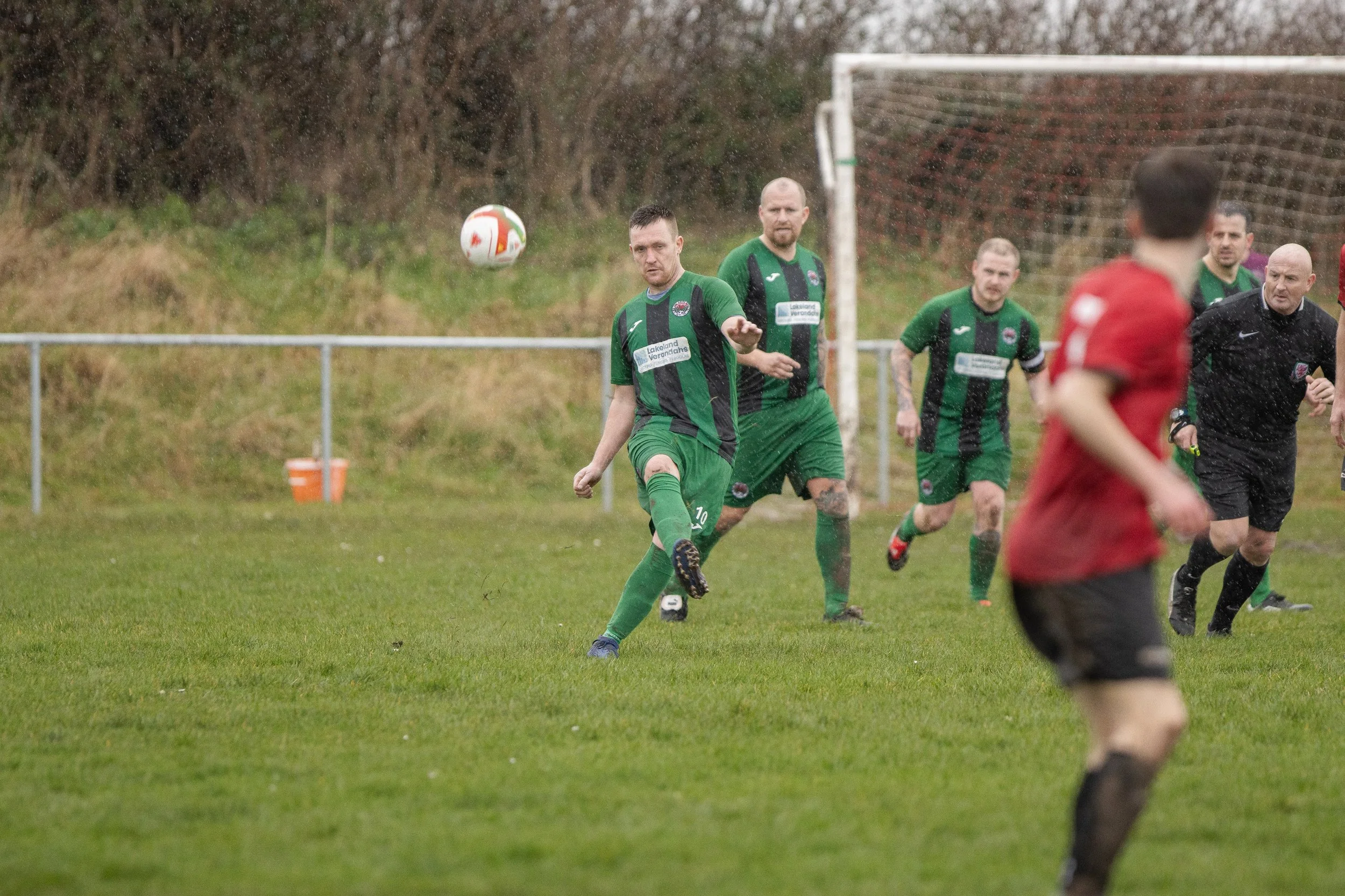 A soccer game in progress during rainy weather, featuring players in green and red jerseys on a grassy field, with a goal in the background.