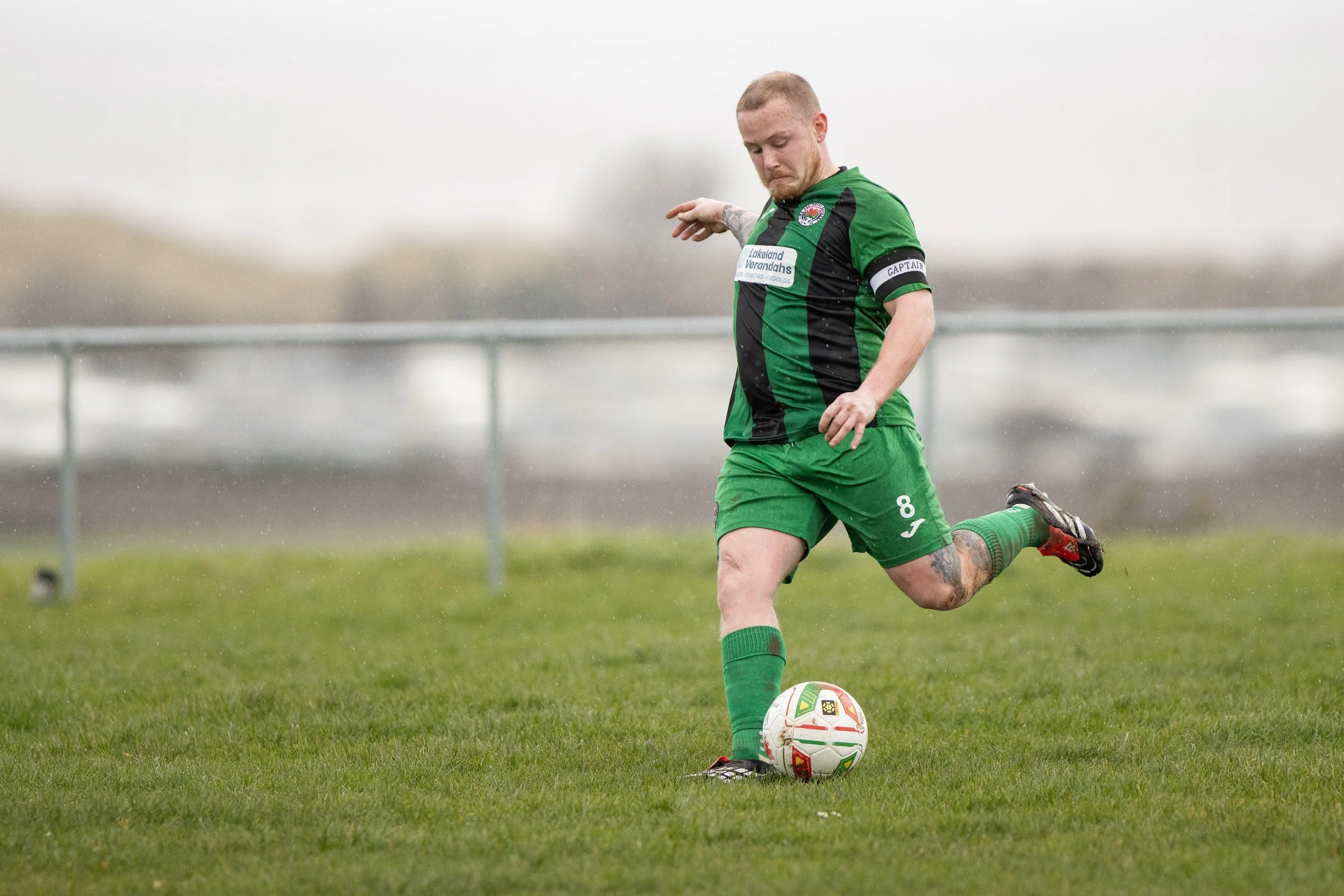 A soccer player in green and black uniform kicks a white soccer ball on a grassy field during a game on a rainy day.