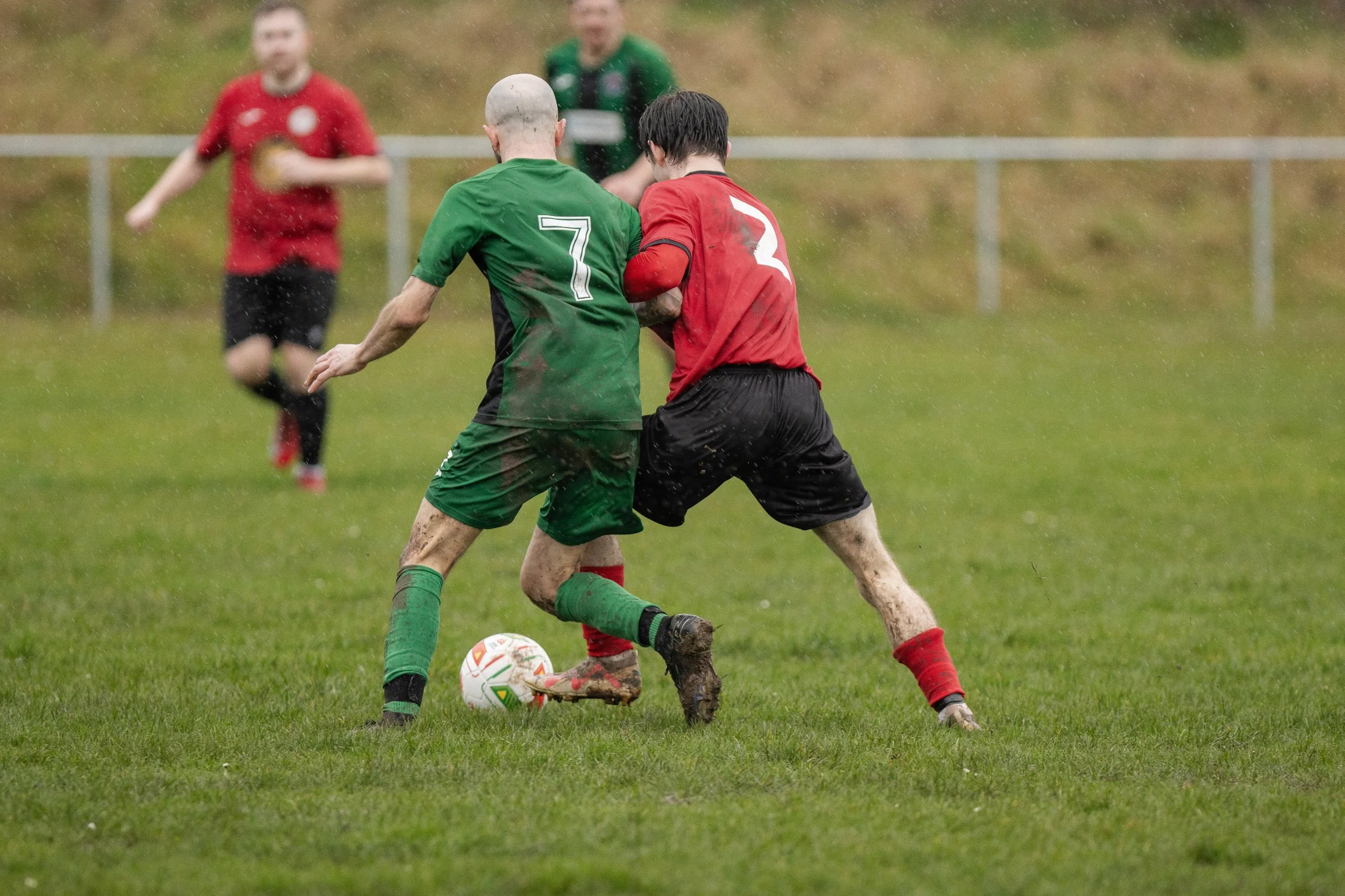 Two soccer players, one in a green uniform with number 7 and one in a red uniform with number 2, are competing for the ball on a wet, grassy field during a game. Other players are visible in the background.