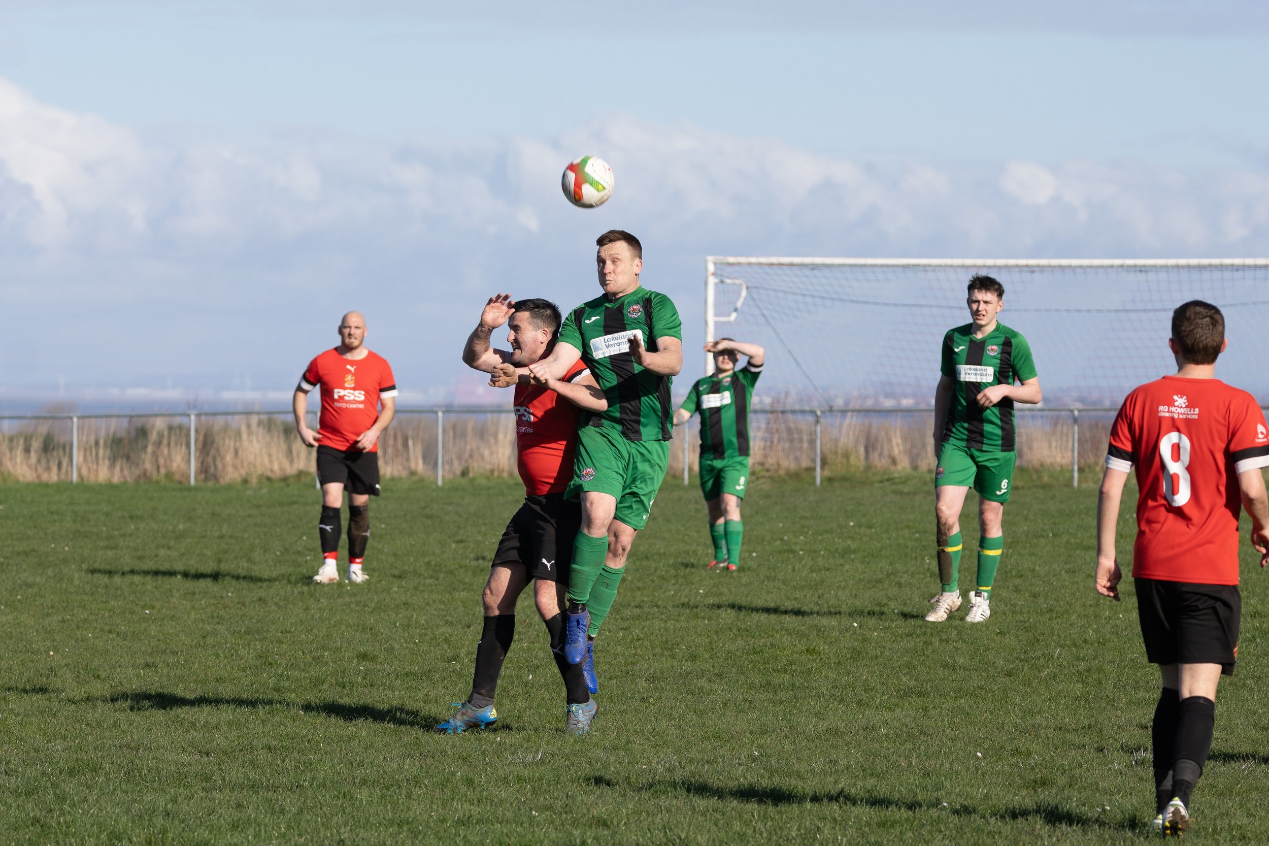 Soccer players on the field competing for a ball in mid-air, with some players in red and others in green uniforms, during a daytime match under a partly cloudy sky.