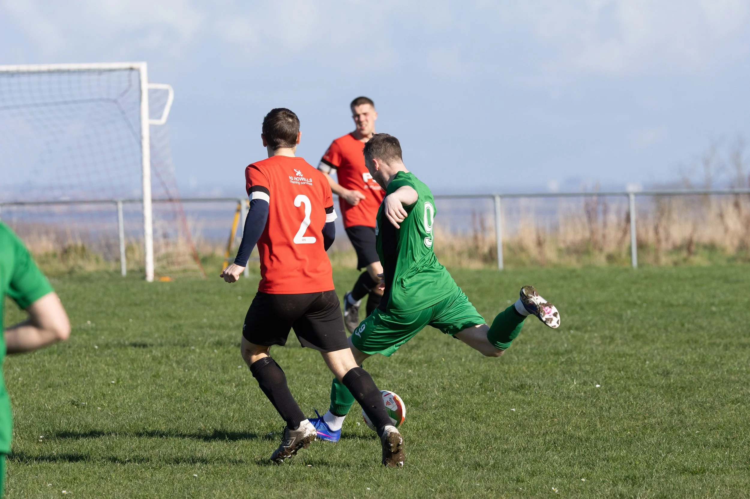 Three soccer players in action on the field, with one in a green jersey kicking the ball while two others in red jerseys attempt to block.